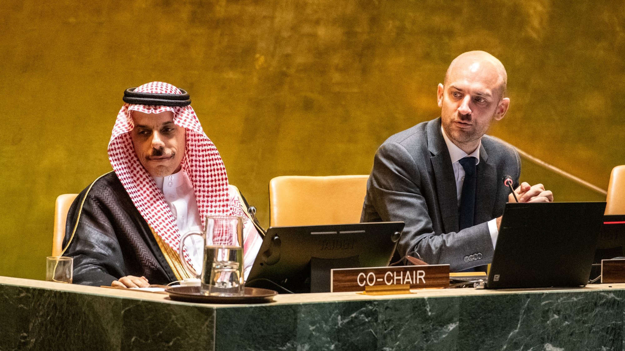 Two men at a conference table, one in traditional attire, discussing on a stage with microphones.