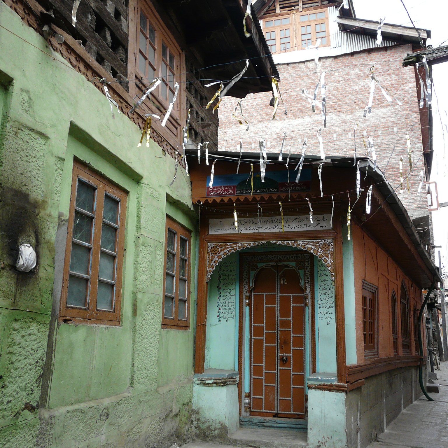 این تصویر نشان‌دهنده یک خیابان باریک در یک منطقه قدیمی است. ساختمان‌ها به سبک سنتی ساخته شده‌اند و رنگ‌های مختلفی دارند. دیوار یک ساختمان به رنگ سبز و دیگری به رنگ نارنجی است که با جزئیات چوبی تزئین شده است. درب چوبی و پنجره‌های با قاب چوبی، زیبایی خاصی به این فضا بخشیده‌اند. درختان و لامپ‌های آویزان نیز جزو دیگر عناصر موجود در تصویر هستند که به فضایی دل‌انگیز و سنتی افزوده‌اند.