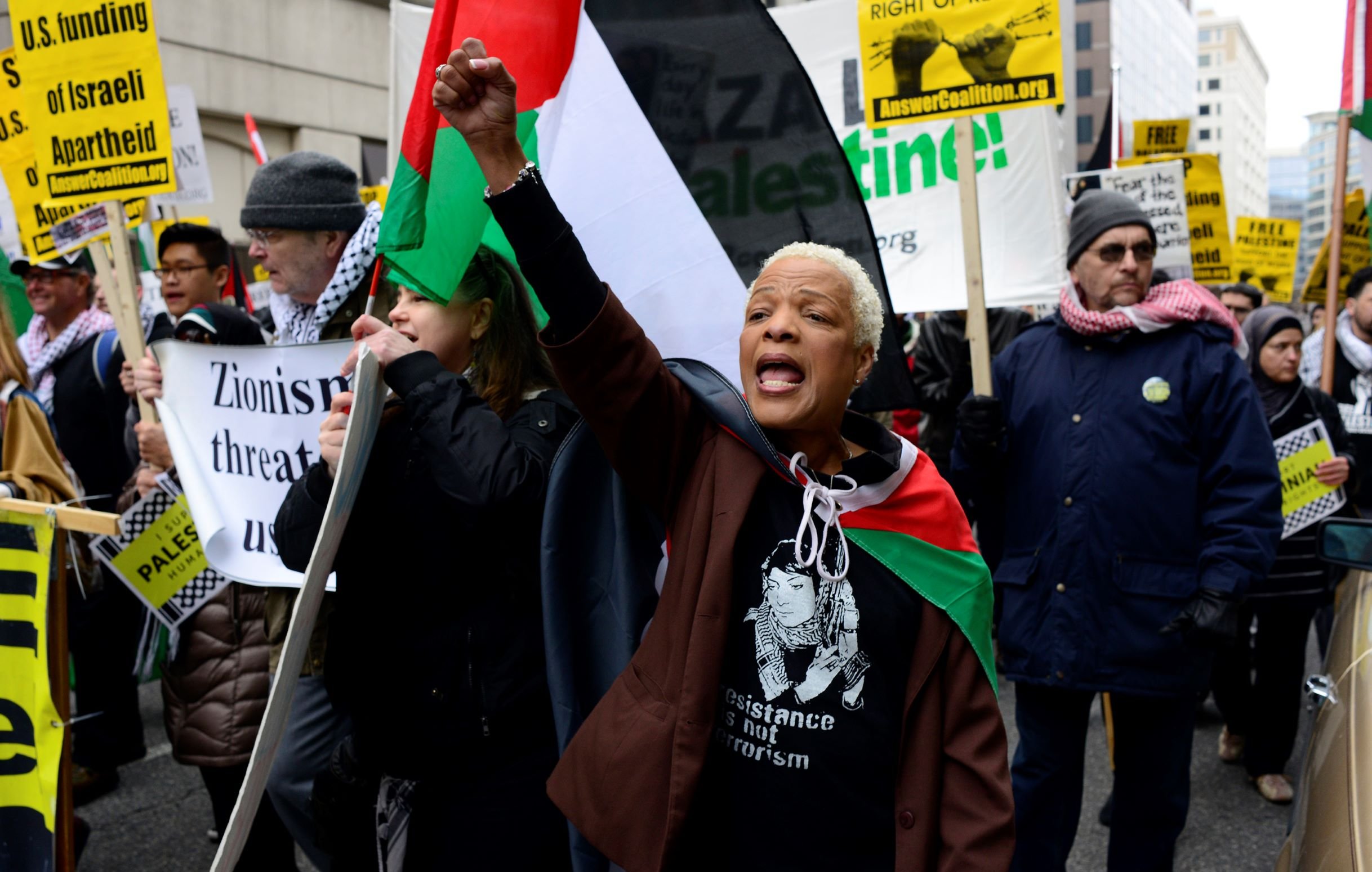 L'image montre une manifestation avec un groupe de personnes brandissant des pancartes et des drapeaux. Une femme au premier plan, portant un t-shirt avec une inscription, lève le poing en criant des slogans. Les manifestants semblent exprimer leur soutien à la cause palestinienne. On peut apercevoir des drapeaux aux couleurs palestiniennes, ainsi que des affiches dénonçant le sionisme et appelant à la résistance. L'ambiance semble dynamique et engagée.
