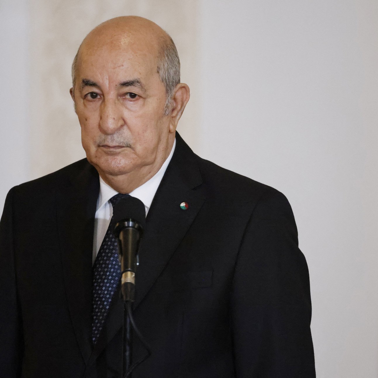The image depicts a middle-aged man dressed in a dark suit, standing at a podium with a microphone. He has a serious expression on his face. In the background, there is an Algerian flag featuring a green and white field with a red star and crescent. The setting appears to be an official environment, likely a government or press conference area.