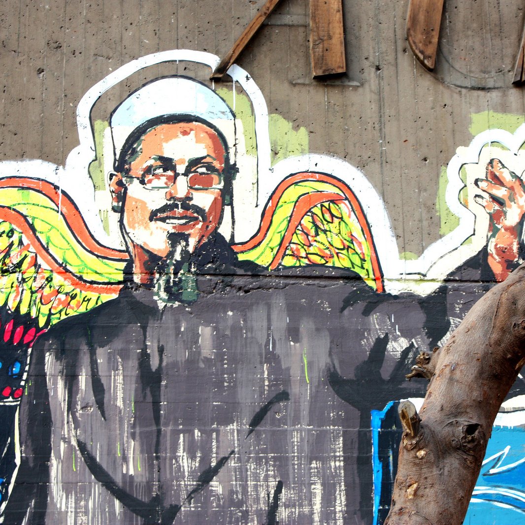 L'image représente une fresque murale colorée. On y voit un homme aux épaules larges, vêtu d'une tunique noire et d'un chapeau traditionnel. Il a un regard expressif et semble faire un geste de la main, comme s'il s'adressait à quelqu'un. En arrière-plan, des motifs vibrants et contrastés, dont des ailes colorées, ajoutent une dimension artistique à la composition. Un arbre, avec des branches dénudées, est également présent au premier plan, soulignant l'environnement urbain de la fresque.