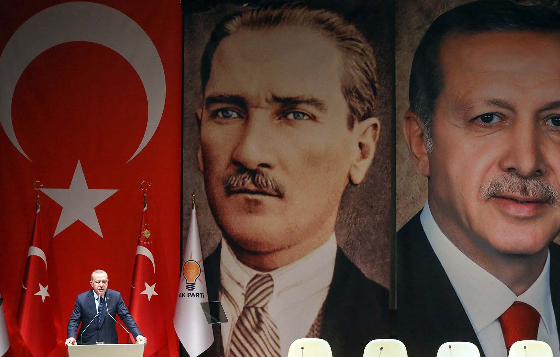 L'immagine ritrae un evento politico in Turchia. Sullo sfondo, sono presenti due grandi ritratti: uno di Mustafa Kemal Atatürk, fondatore della Repubblica turca, e l'altro di Recep Tayyip Erdoğan, attuale presidente. Al centro, un oratore sembra parlare al pubblico, con bandiere turche e simboli del partito sul palco. Lo sfondo con i due leader storici suggerisce un legame tra il passato e il presente politico del paese.