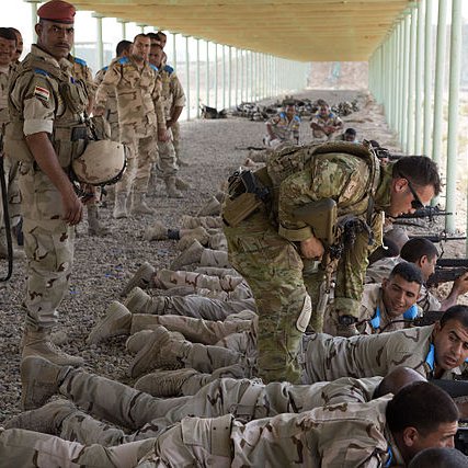 L'image montre un groupe de soldats en train de s'entraîner sur un champ de tir, allongés à terre avec leurs armes. Certains soldats portent des uniformes beiges avec des motifs camouflés, tandis qu'un instructeur semble donner des conseils à un autre soldat. En arrière-plan, on aperçoit d'autres soldats attendant ou surveillant l'exercice. L'environnement est désertique avec une structure en métal au-dessus d'eux, offrant un abri contre le soleil.
