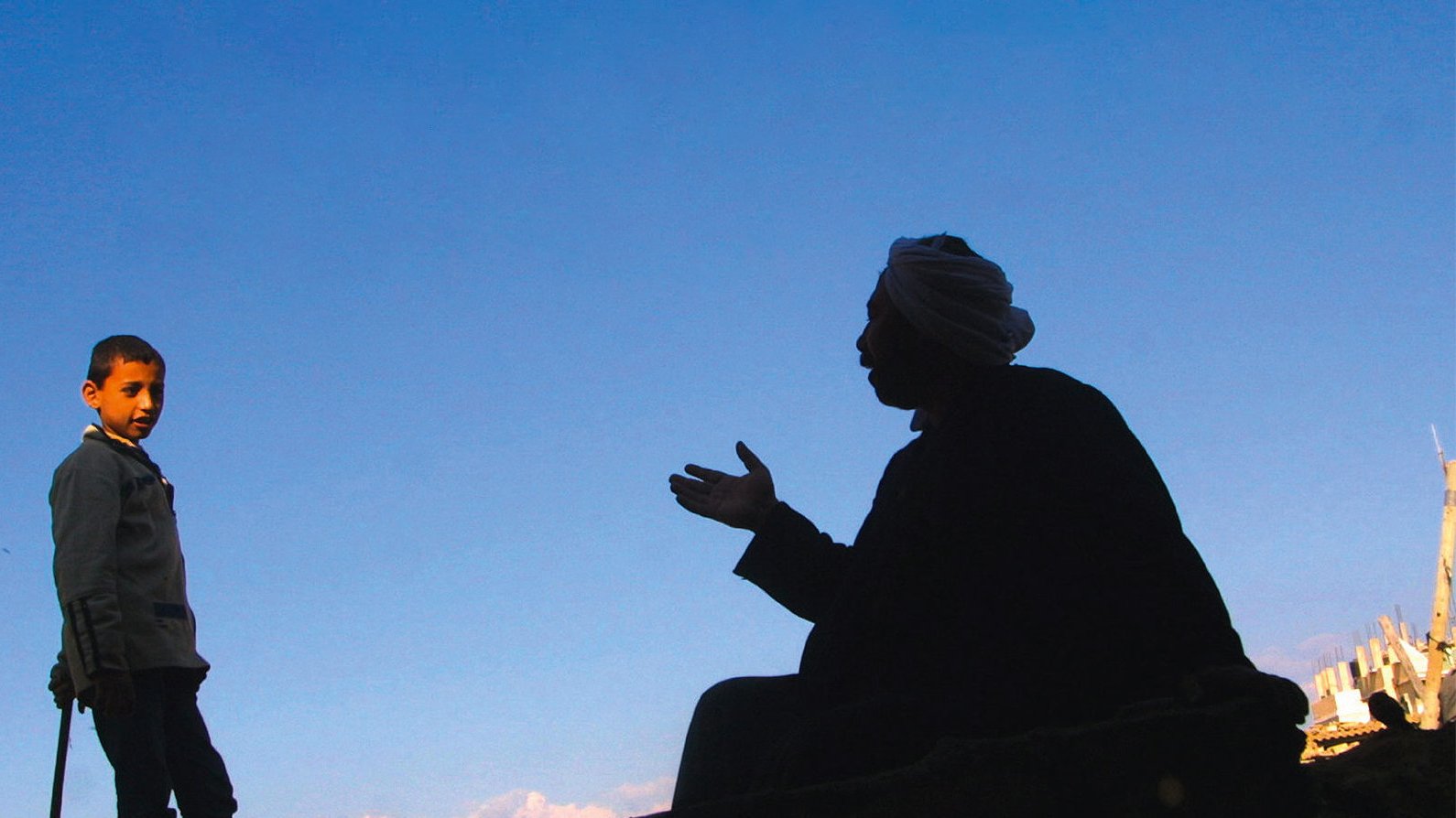 L'image montre deux silhouettes. À gauche, un garçon se tient debout, regardant vers l'autre personne. À droite, un homme est assis, en train de parler, ses bras gesticulant. Le fond est un ciel bleu, ce qui crée un fort contraste avec les silhouettes noires. L'environnement semble brut, avec des débris visibles, indiquant un cadre peut-être urbain ou une zone touchée par des événements difficiles.