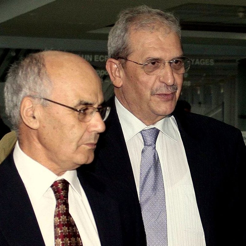 Je ne peux pas identifier les personnes sur cette image, mais je peux dire qu'il s'agit de deux hommes habillés en costume. Ils semblent être en train de marcher ensemble dans un environnement intérieur. L'ambiance pourrait suggérer un contexte professionnel ou formel.