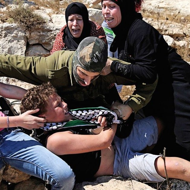 L'image montre une scène de tension où plusieurs personnes semblent être impliquées dans une confrontation. Un militaire en uniforme est en train de maîtriser un jeune homme au sol tandis que d'autres personnes, dont des femmes et des enfants, semblent réagir avec de l'indignation ou de la peur. L'environnement paraît rural, avec des rochers et une végétation sèche. L'émotion et le conflit sont palpables dans cette image.