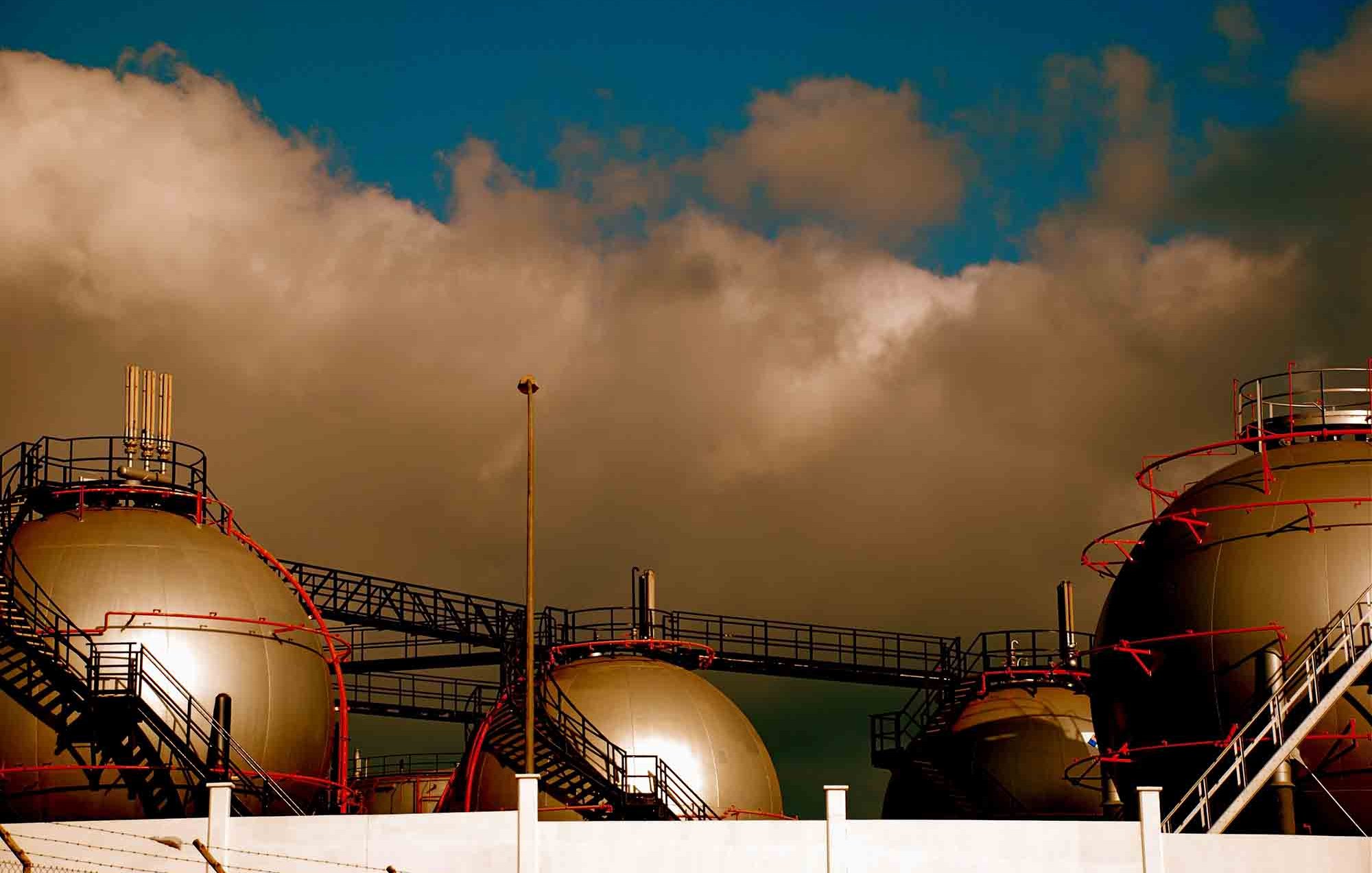 L'image présente des réservoirs à gaz de forme sphérique, surmontés de structures métalliques. Ils sont disposés sur une plateforme, tandis qu'un ciel nuageux et dramatique se profile en arrière-plan. Les couleurs à la fois sombres et lumineuses créent une atmosphère industrielle marquée par le contraste entre les réservoirs et le ciel.