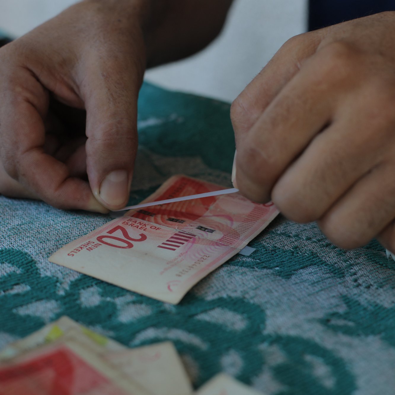 L'immagine mostra delle mani che stanno piegando e preparando delle banconote, probabilmente di grosso taglio. Le banconote sono adagiate su un tavolo con una superficie di tessuto verde, e accanto si vede un rotolo di nastro adesivo. I movimenti delle mani suggeriscono che si sta cercando di raggruppare o sistemare il denaro in modo ordinato.