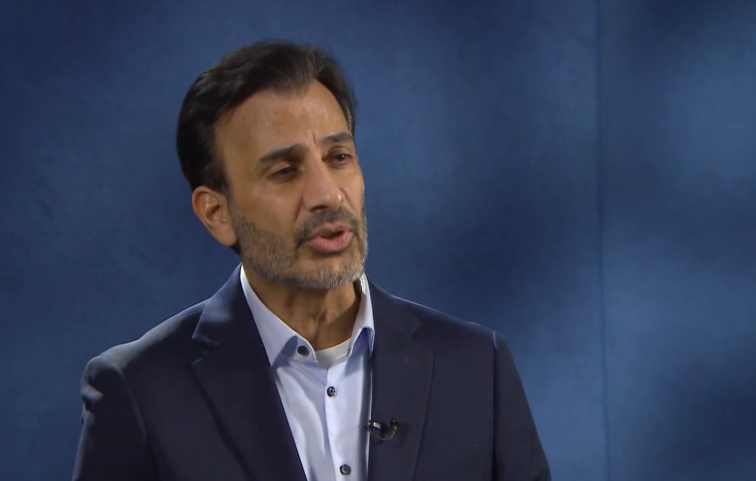 Nell'immagine si vede un uomo con una barba curata e capelli scuri, vestito con una giacca scura e una camicia chiara. Sembra che stia partecipando a un'intervista o a un discorso, mentre si esprime con attenzione. Sullo sfondo c'è un'ambientazione di colore blu, che crea un'atmosfera professionale.