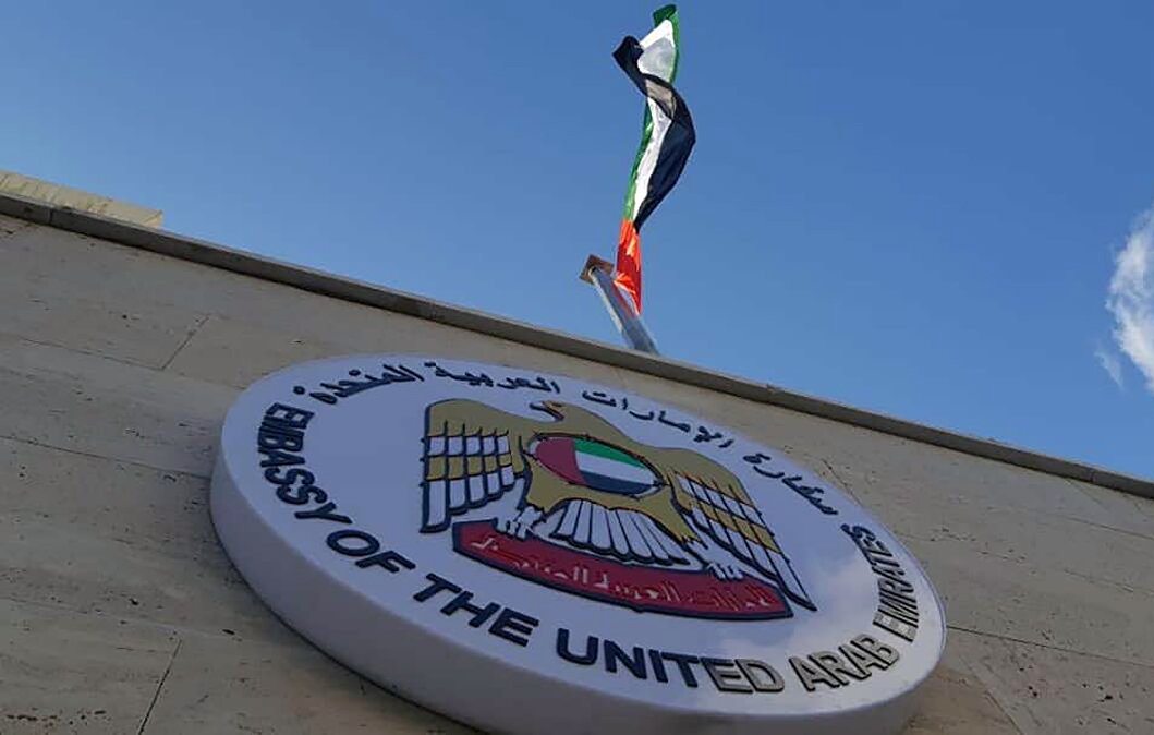 این تصویر نمایی از سفارت امارات متحده عربی را نشان می‌دهد. در بالای ساختمان، پرچم امارات به اهتزاز درآمده است. در پایین تصویر، علامت یا نشان سفارت با نوشته‌ای به زبان انگلیسی "EMBASSY OF THE UNITED ARAB EMIRATES" به چشم می‌خورد. آسمان آبی و ابرهایی نیز در پس‌زمینه مشاهده می‌شود.