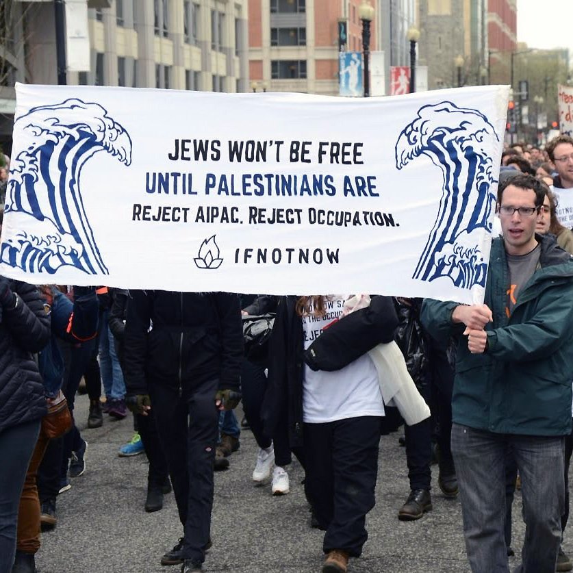 The image shows a group of protesters marching together, holding a large banner that reads, "Jews won't be free until Palestinians are. Reject AIPAC. Reject occupation." The banner features artistic waves, and the participants appear to be actively engaged in the demonstration, dressed in casual winter clothing. The background includes urban buildings and other protesters. The atmosphere conveys a sense of solidarity and activism focused on social justice issues.