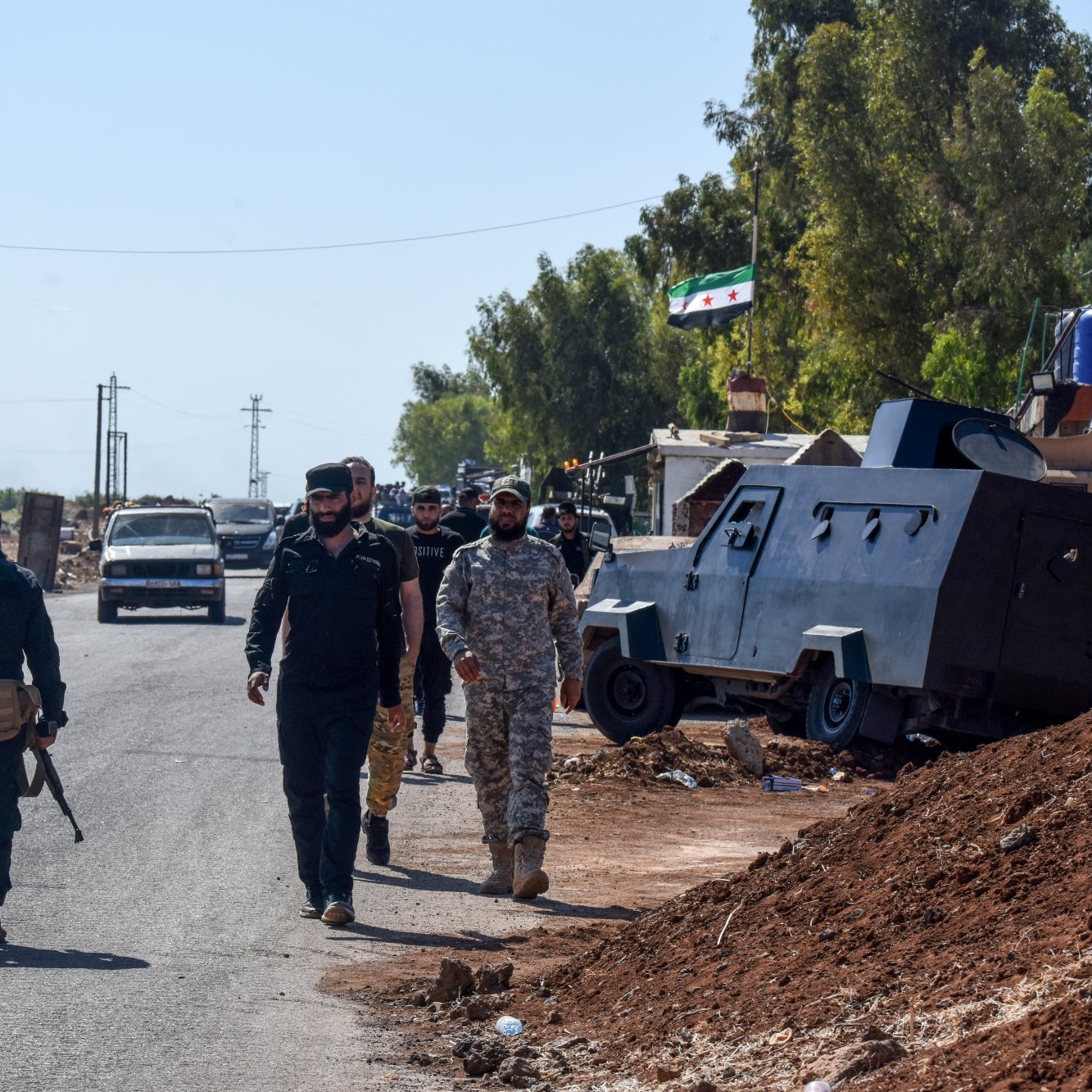 تصویر، یک خیابان با خودروها و کامیون‌ها است. چند نفر با لباس نظامی در حال حرکت هستند. دیوارها و درختان اطراف هستند.