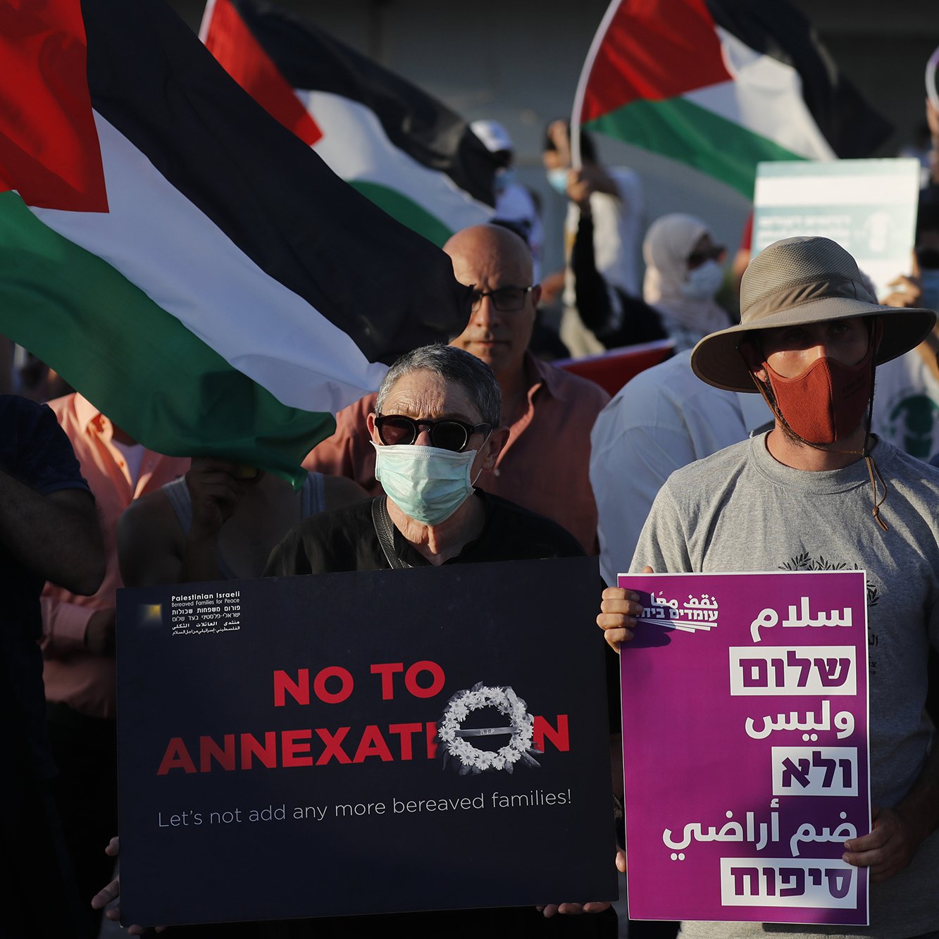 The image depicts a group of protesters holding flags and signs during a demonstration. The flags feature the colors of the Palestinian flag. Some participants are wearing masks, and a few signs read messages against annexation and calls for peace. The atmosphere appears to be one of solidarity and activism, as the individuals gather for a common cause.