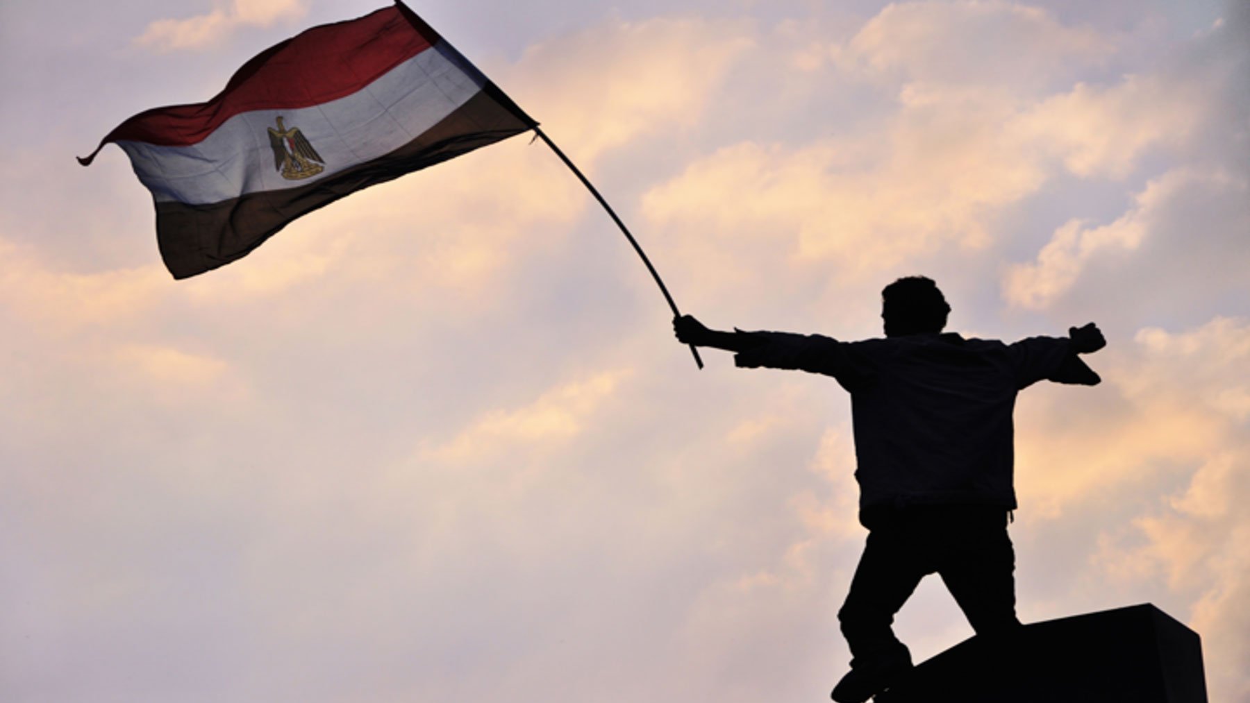 L'image montre une silhouette d'une personne debout sur une plateforme, levant un drapeau. Le drapeau est égyptien, avec des couleurs rouge, blanc, noir et le symbole du pays. L'arrière-plan présente un ciel avec des nuages aux teintes douces, créant une ambiance de détermination et de célébration. Cette scène évoque des sentiments de patriotisme et de protestation ou de victoire.