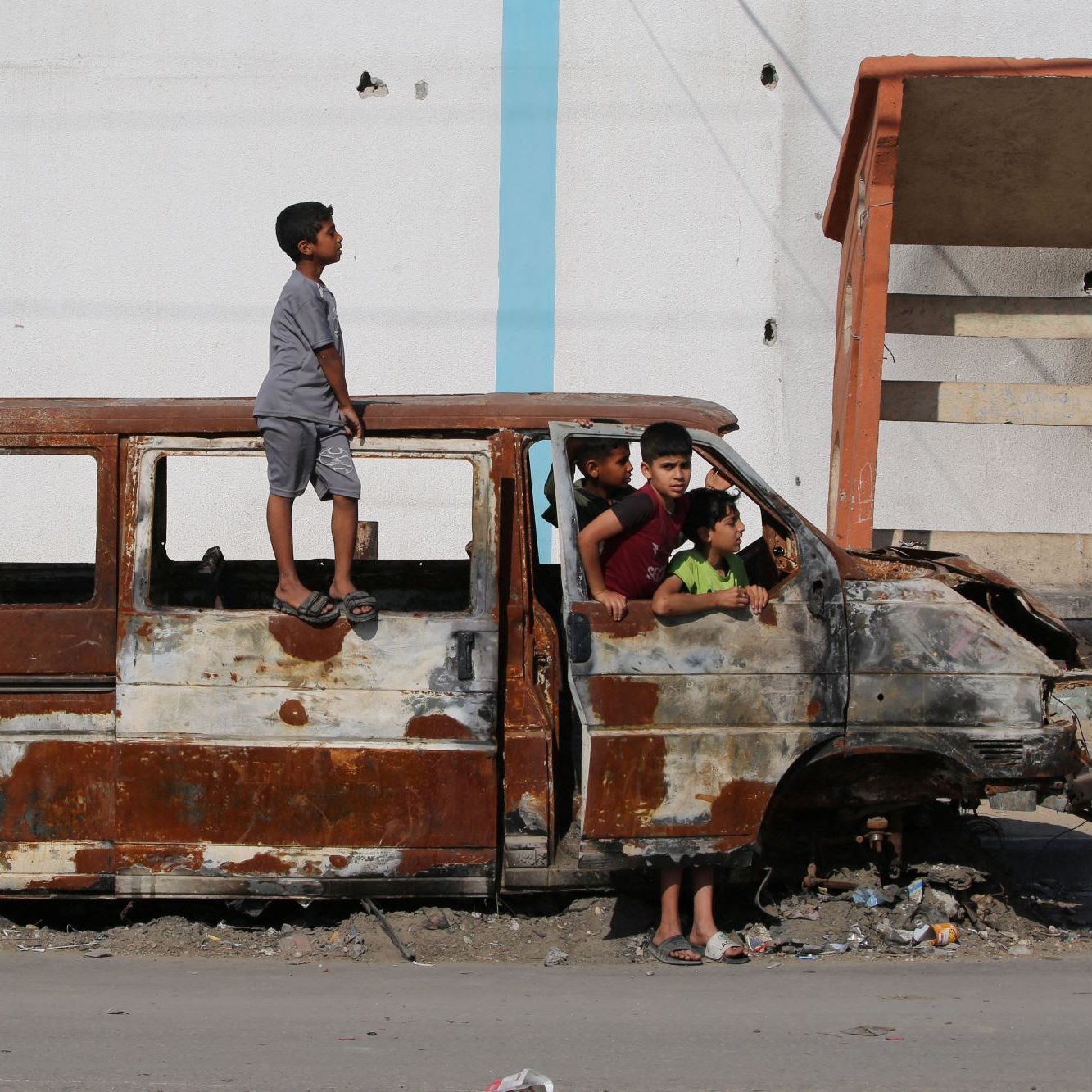 در این تصویر، یک خودرو قدیمی و زنگ‌زده دیده می‌شود که در یک محیط شهری و آشفته قرار دارد. چند کودک در اطراف این خودرو بازی می‌کنند و یک کودک بر روی سقف آن ایستاده است. مناظر پشت سر آن‌ها شامل دیوارهای آسیب‌دیده و ناپایداری‌های دیگر است که نشان‌دهنده شرایط سخت و فرسوده منطقه است. این تصویر احساساتی از کودکی و بازی در شرایط نامناسب را منتقل می‌کند.