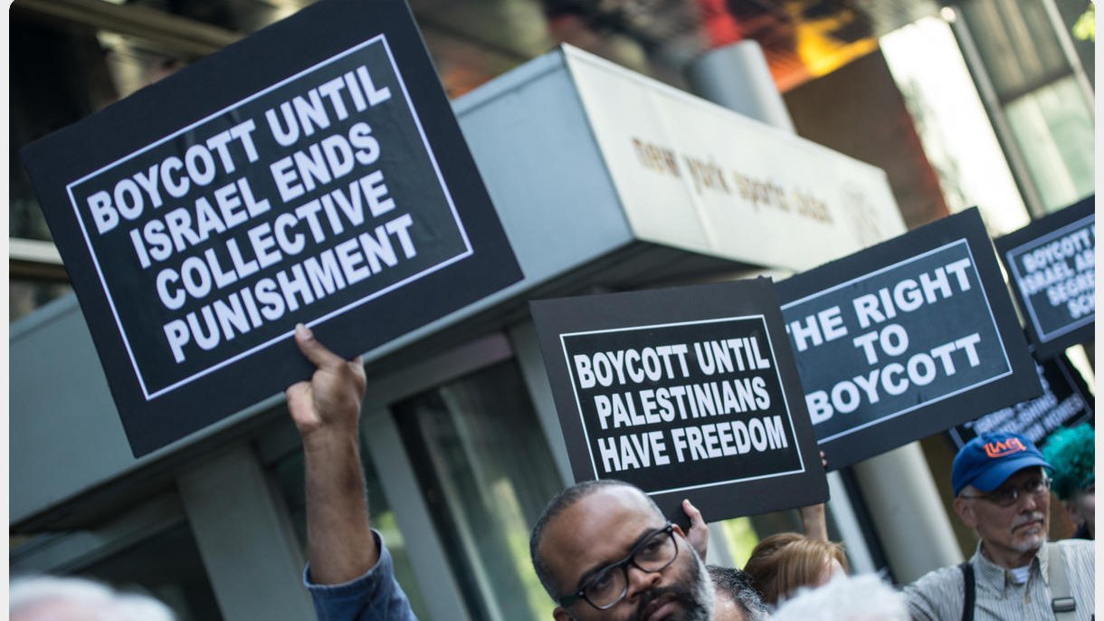 The image depicts a group of protesters holding signs at a demonstration. Some of the signs read "BOYCOTT UNTIL ISRAEL ENDS COLLECTIVE PUNISHMENT" and "BOYCOTT UNTIL PALESTINIANS HAVE FREEDOM." The atmosphere suggests a call for social justice and awareness regarding the Palestinian situation. The protesters appear engaged and determined in their stance.