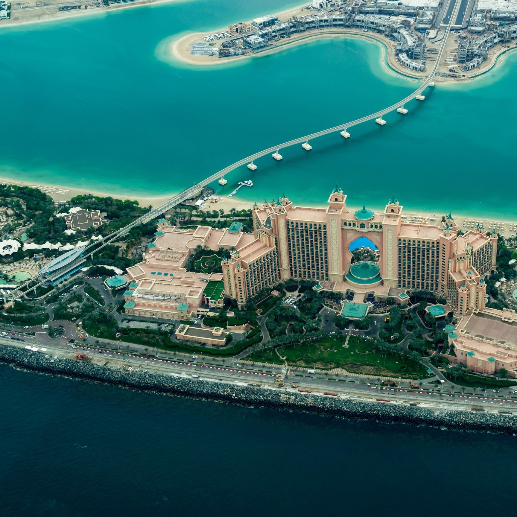 L'image montre une vue aérienne d'un impressionnant complexe hôtelier situé sur le bord de la mer. On peut voir un grand bâtiment aux couleurs beige et rose, entouré de jardins bien entretenus et de plusieurs piscines. La mer turquoise qui l'entoure contraste avec les structures urbaines modernes visibles en arrière-plan. Un pont relie le complexe à une autre zone, et l'ensemble est à la fois luxueux et captivant, évoquant une destination touristique prisée.