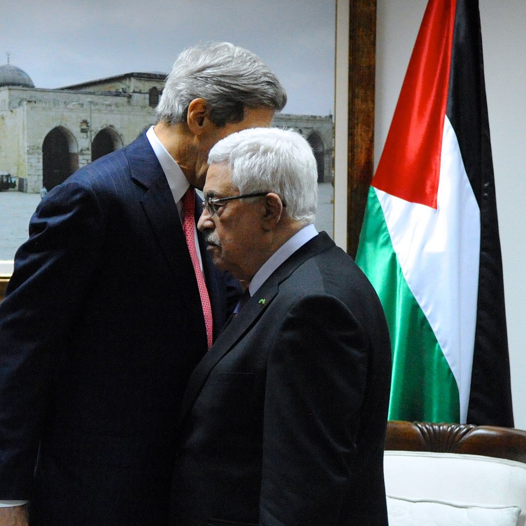 L'image montre deux hommes en train de s'incliner l'un vers l'autre dans un environnement formel. En arrière-plan, il y a une grande photo d'un monument architectural, probablement une mosquée, et un drapeau représentant les couleurs rouge, noir, blanc et vert. Les hommes semblent avoir une conversation sérieuse, et l'un d'eux est habillé en costume, tandis que l'autre porte une tenue plus traditionnelle. L'atmosphère de la scène évoque un moment de négociation ou de discussion diplomatique.