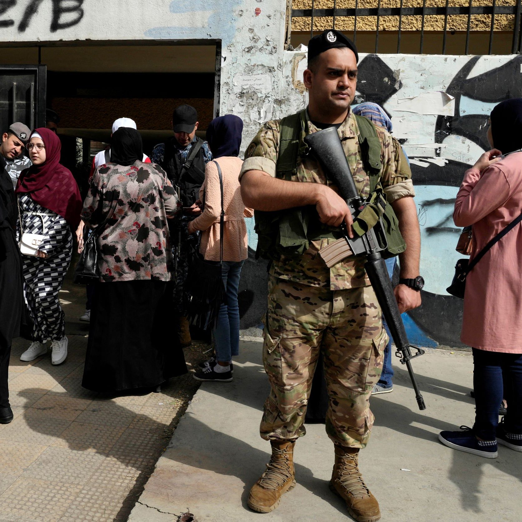 La imagen muestra a un soldado de pie con un rifle, vigilando una entrada. Al fondo, hay un grupo de mujeres, algunas de las cuales llevan hijab, esperando. La escena parece tener lugar en un ambiente urbano, con murales o grafitis en las paredes cercanas. El soldado parece atento y en posición de guardia, mientras las mujeres se agrupan en la entrada.
