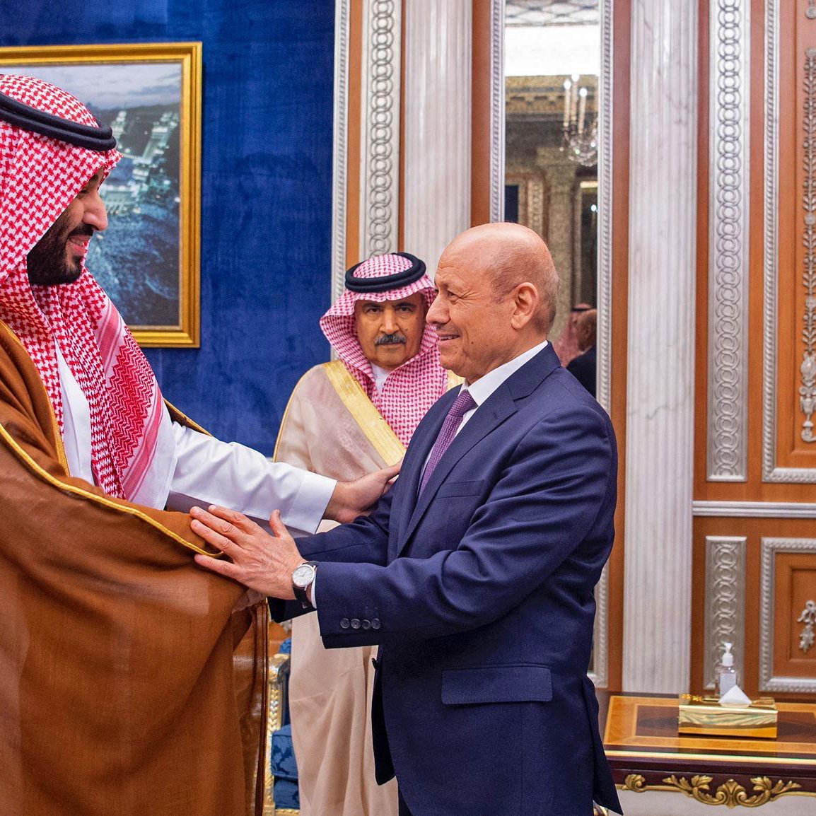 در این تصویر، دو مرد در حال گفت‌وگو و خوش‌آمدگویی به یکدیگر هستند. یکی از آن‌ها لباس سنتی عربی به نام "ثوب" به تن دارد و چفیه‌ای خاص بر سر دارد. او به نظر می‌رسد که در مقام رهبری قرار دارد. مرد دیگر نیز به صورت رسمی لباس پوشیده است و نشان از احترام و ادب در بیان حرکات او دیده می‌شود. در پس‌زمینه، دکوراسیون مجلل و لوکس با تزئینات طلایی و تابلوهایی از مناظر شهری دیده می‌شود که به فضای رسمی این ملاقات افزوده است.