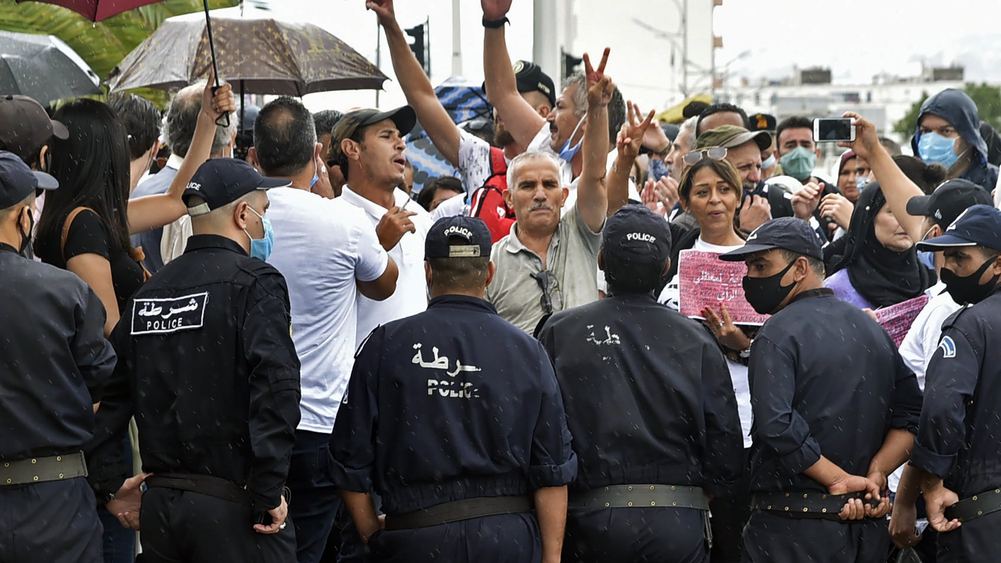 L'image montre une scène de tension entre un groupe de manifestants et des policiers. Les manifestants, certains avec des parapluies, lèvent les bras et expriment leurs opinions. Les policiers, en uniforme et portant des masques, se tiennent en face d'eux, créant un contraste clair entre les deux groupes. La situation semble chargée d'émotion, avec des personnes qui cherchent à faire entendre leur voix. Le temps est pluvieux, ajoutant une atmosphère dramatique à l'événement.