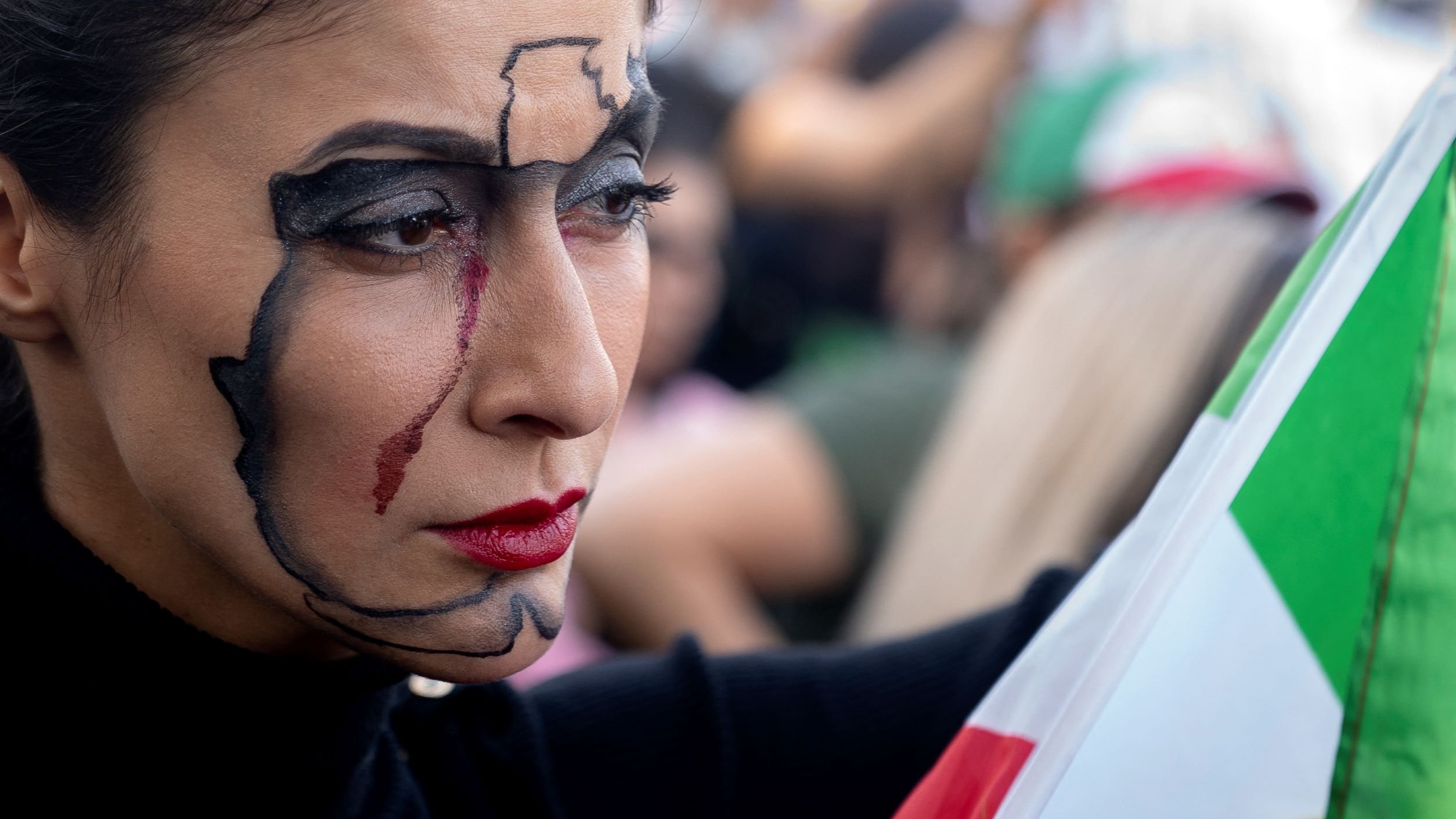 این تصویر زنی را نشان می‌دهد که در یک تجمع عمومی حضور دارد. او صورت خود را با طراحی خاصی تزئین کرده است که نقشه‌ای از یک کشور یا منطقه را به تصویر می‌کشد و در کنار آن اثراتی از خط سیاه و رد خون مشاهده می‌شود. او همچنین لب‌های قرمز و پوشش تیره به تن دارد و در دستش پرچمی با رنگ‌های مختلف را نگه داشته است. چهره‌اش نشان‌دهنده‌ی احساس جدی و تأمل است. این تصویر به نظر می‌رسد به یک حرکت اجتماعی یا سیاسی مربوط باشد.