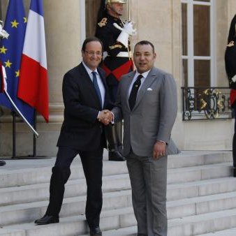 L'image montre une scène officielle à l'extérieur d'un bâtiment gouvernemental. Deux hommes, en costume, se serrent la main sur des marches, symbolisant une rencontre diplomatique. À l'arrière-plan, on peut voir des drapeaux de la France et du Maroc, ainsi que des soldats en tenue d'apparat. L'atmosphère est formelle et solennelle, reflétant l'importance de l'événement.