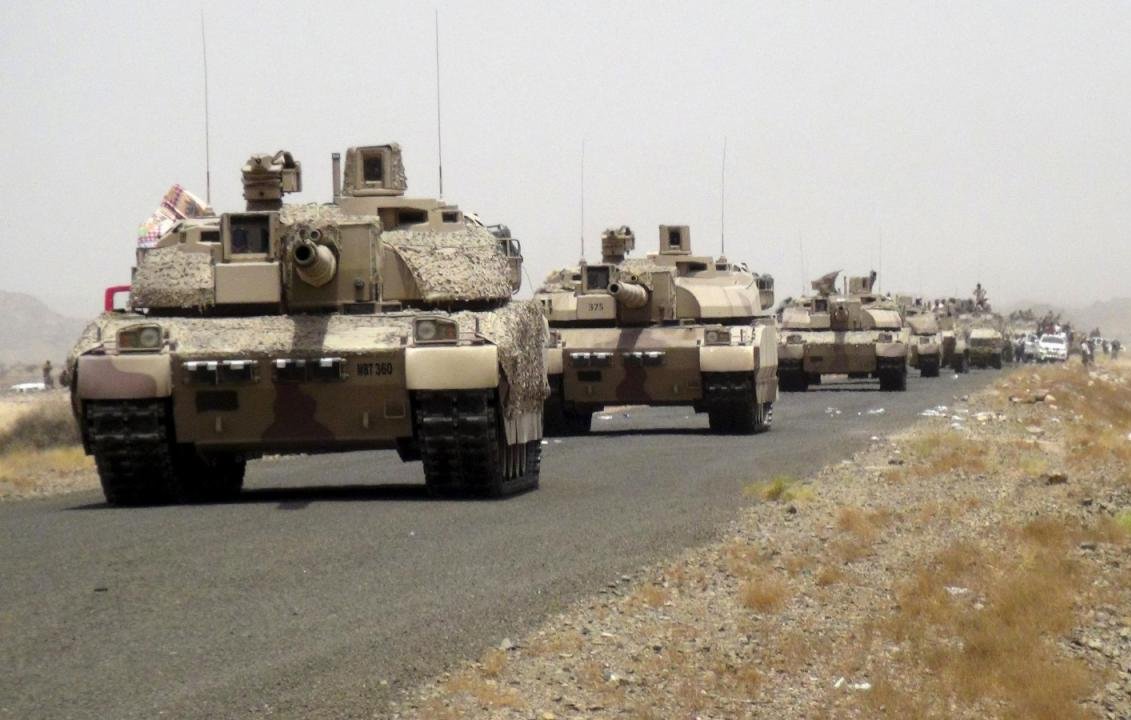 تظهر الصورة قافلة من الدبابات العسكرية تسير على طريق ترابي. يبدو أن المركبات مموهة بألوان تتماشى مع البيئة الصحراوية. في الخلفية، يمكن رؤية جبال أو تلال ذات لون بني محايد. الأجواء تبدو غائمة بعض الشيء، مما يشير إلى وجود غبار أو ضباب خفيف. الدبابات مرتبة بشكل منظم، مما يدل على تحركها كجزء من عملية عسكرية أو تمرين.