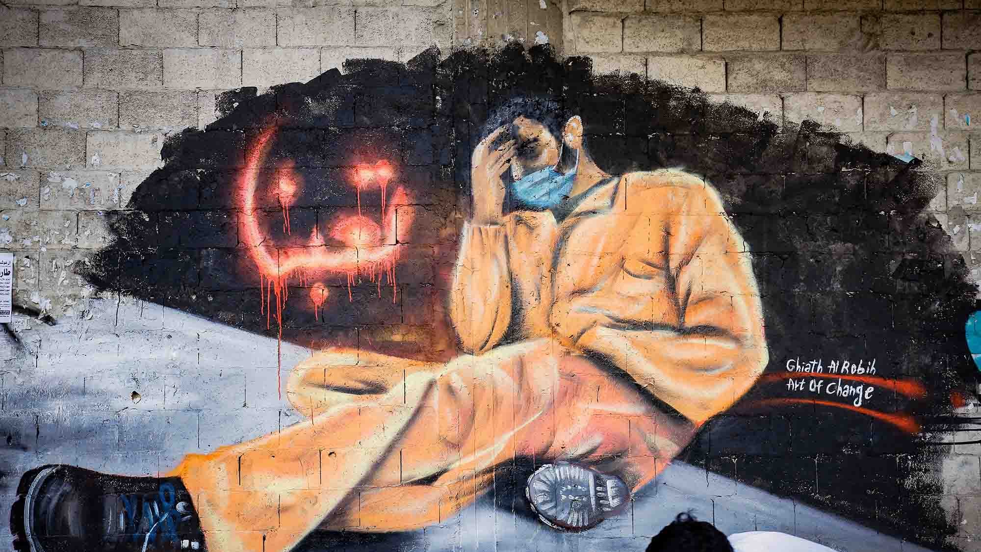 L'immagine mostra un murales che rappresenta una figura umana seduta, vestita con una tuta arancione, che tiene la testa tra le mani in un gesto di disperazione o riflessione. Il personaggio sembra essere in una posizione di vulnerabilità e solitudine. Accanto a lui, ci sono scritte in arabo e gocce di colore rosso che aggiungono un senso di drammaticità. Nella parte inferiore della scena, si vede un ragazzo in posizione inginocchiata, che sembra osservare il murales. Questo opera sembra voler trasmettere un messaggio profondo sulla sofferenza e l'oppressione.