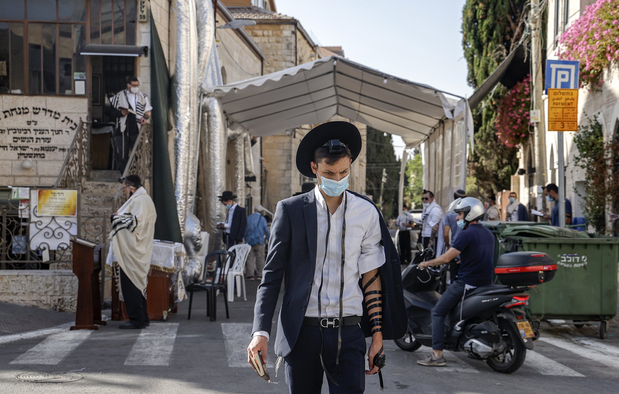En la imagen se puede ver a un hombre joven que lleva puesta una camisa blanca y un saco oscuro, además de un sombrero negro. Está caminando por una calle que parece estar cerca de un lugar de culto judío, ya que al fondo se observa un edificio con elementos relacionados con la cultura y religión judía. El hombre lleva una máscara, posiblemente debido a las precauciones sanitarias. Se puede notar que tiene filacterias (tefilín) en su brazo, lo cual es parte de la práctica judía durante las oraciones. A su alrededor hay otras personas, algunas en la acera y otras en vehículos, lo que sugiere que es un lugar activo. Hay también un toldo montado, probablemente utilizado para eventos religiosos.