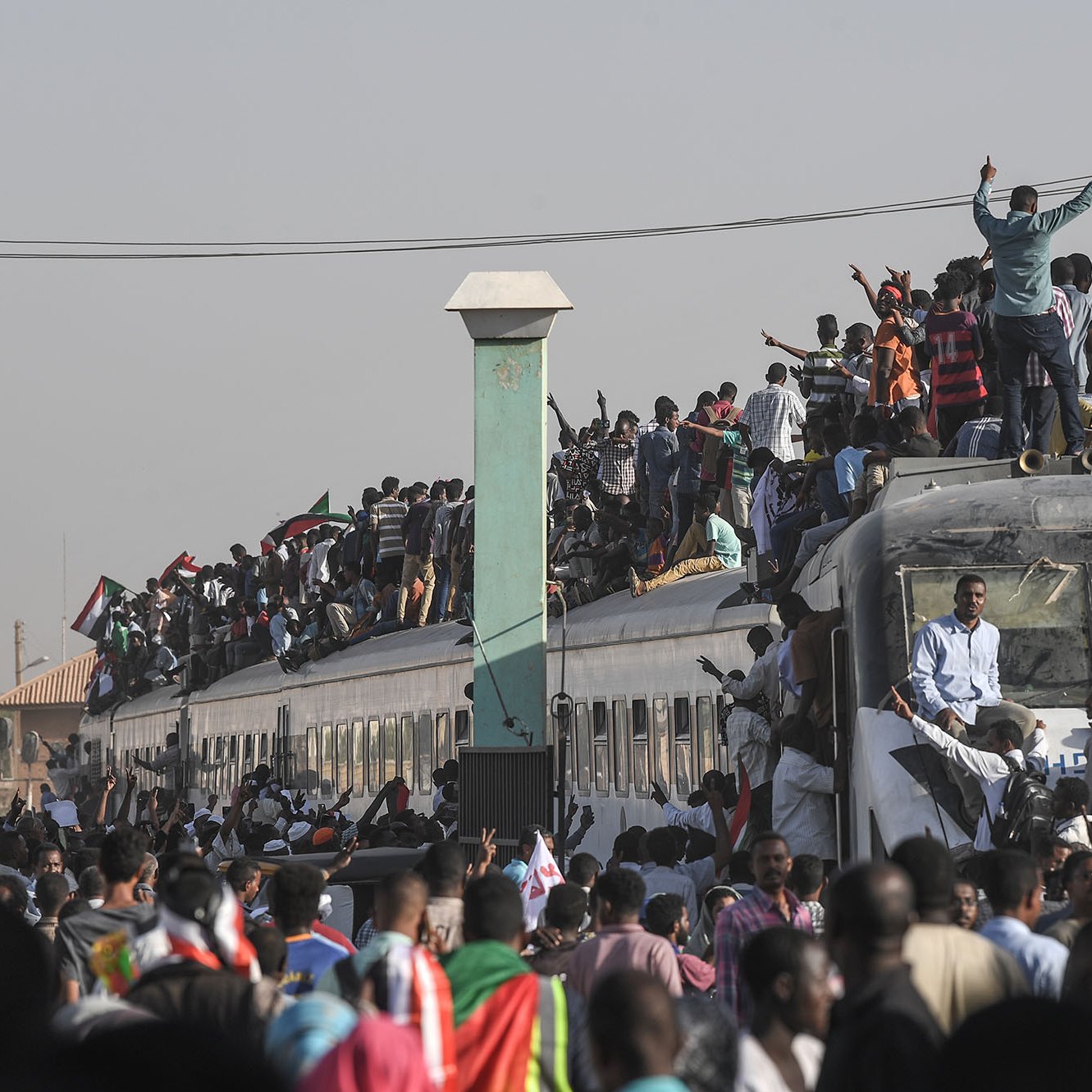 L'image montre une grande foule de personnes rassemblées autour et sur un train. Beaucoup de gens se tiennent sur le toit du train, tandis que d'autres s'accrochent aux côtés. L'atmosphère semble vibrante et énergique, probablement liée à une manifestation ou un événement important. En arrière-plan, on peut apercevoir des bâtiments et des infrastructures typiques d'une ville. La scène dégage un sentiment de solidarité et de détermination parmi les participants.