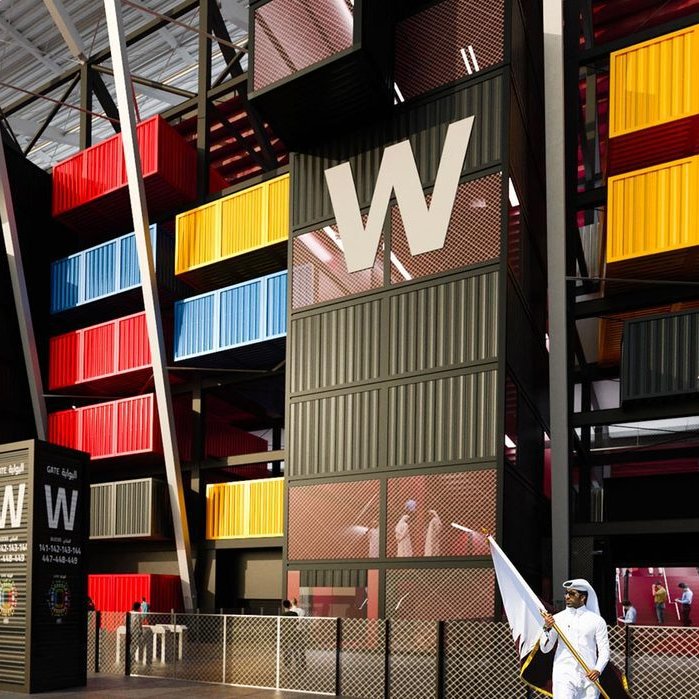 The image depicts a modern architectural structure that resembles a stadium or a large public venue. The building features a striking façade made up of brightly colored shipping containers in red, yellow, and blue, arranged in a visually dynamic way. There are tall, angular elements with a white roof framework above. In the foreground, a group of people dressed in traditional attire can be seen, indicating a cultural context. Overall, the design conveys a sense of contemporary style and innovative use of space.