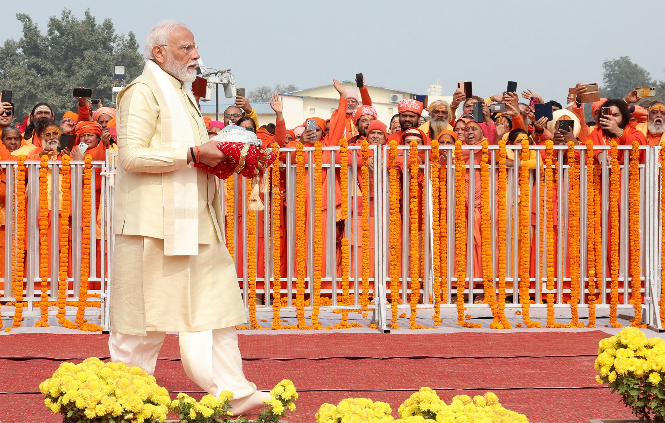 En la imagen se puede ver a un hombre con barba y cabello canoso vestido con un traje tradicional claro, que sostiene un objeto en sus manos. Este hombre camina hacia la derecha, mientras que detrás de él hay un grupo de personas vistiendo túnicas naranjas. Están emocionadas y algunas de ellas levantan sus teléfonos móviles para capturar el momento. La escena está decorada con flores amarillas y hay una valla blanca que separa al hombre de la multitud. En el fondo, se observa un ambiente festivo y colorido.