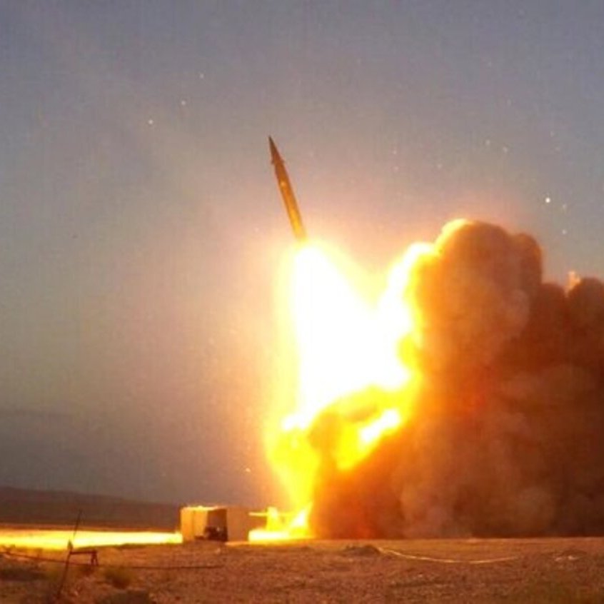 L'immagine mostra un razzo lanciato da un sistema di lancio mobile. Si può notare un intenso getto di fiamme e fumi all'uscita del razzo, accompagnato da una nube di polvere sollevata dal terreno. Il cielo è visibile sullo sfondo, indicando un momento di sera o al crepuscolo, con tonalità che suggeriscono l'imminente oscurità. La scena trasmette una forte impressione di potenza e dinamismo.