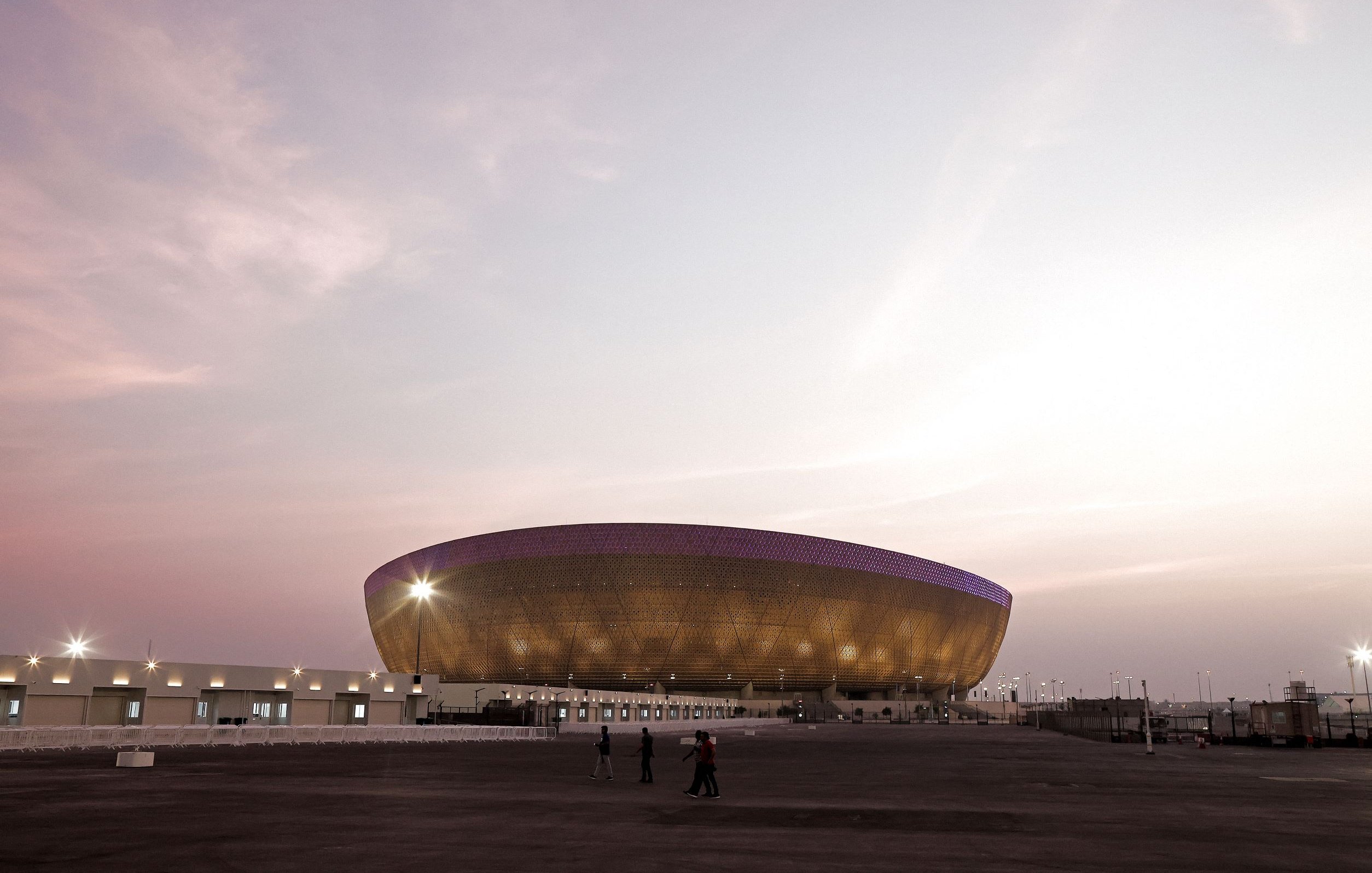 The image shows a large, modern stadium with a unique, oval design. The stadium features an exterior of golden-hued materials, giving it a striking appearance, especially during twilight. There are a few people walking in the foreground, and the sky is painted in soft pastel colors, suggesting either dawn or dusk. The stadium is well-lit, with lights illuminating its structure and the surrounding area. The overall atmosphere is calm and serene, with a sense of anticipation for an event.