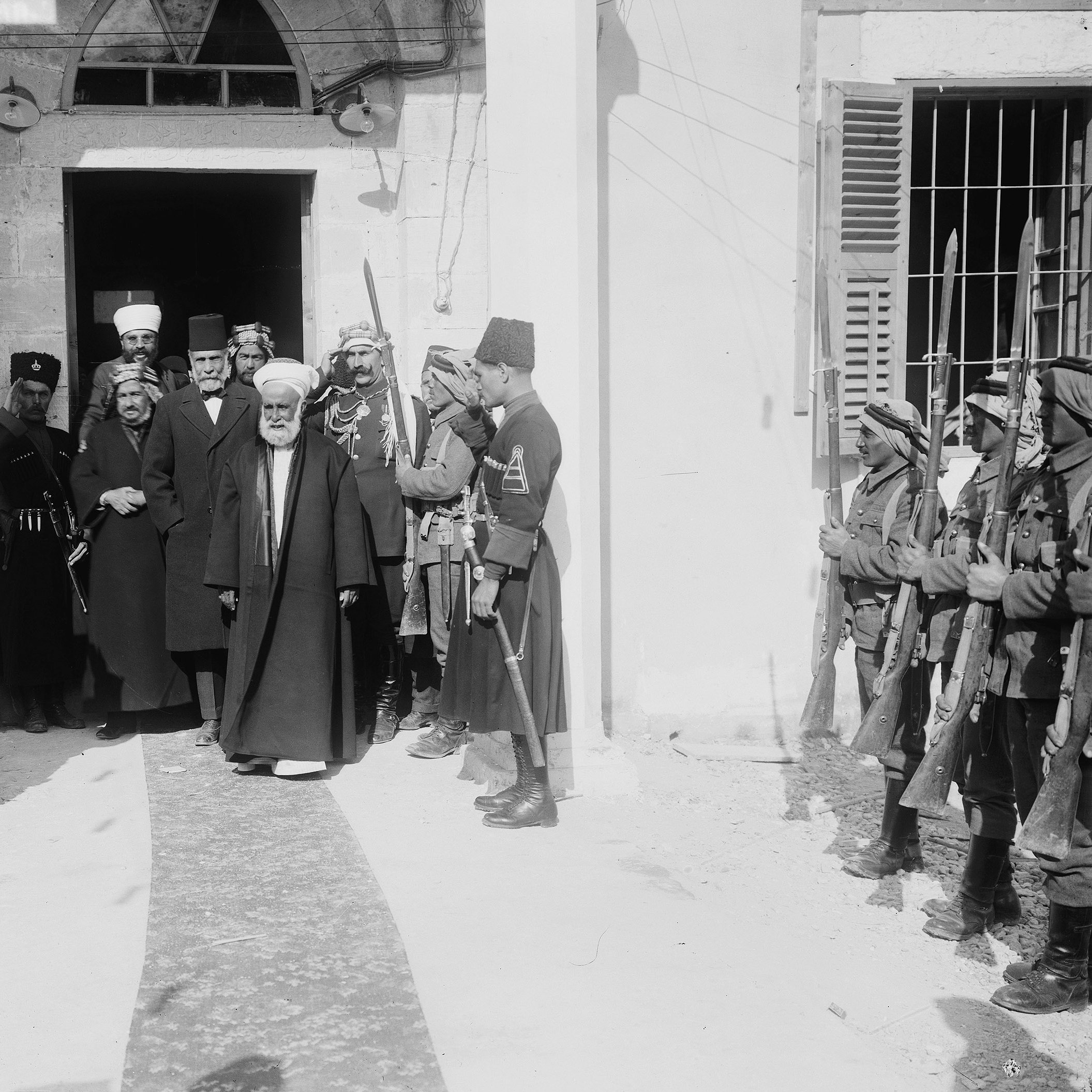 در تصویر، گروهی از مردان در یک مکان رسمی ایستاده‌اند. چند نفر از آنها در لباس‌های نظامی و دیگران در لباس‌های سنتی دیده می‌شوند. مردی با چهره‌ای جدی و کت و شلوار تیره در حال قدم گذاشتن به سمت دوربین است، در حالی که دیگران به او احترام می‌گذارند و سلام نظامی می‌دهند. محیط تصویر به نظر می‌رسد که یک ساختمان تاریخی یا دولتی باشد. فضای کلی نشان‌دهنده‌ی یک واقعه مهم و رسمی است.