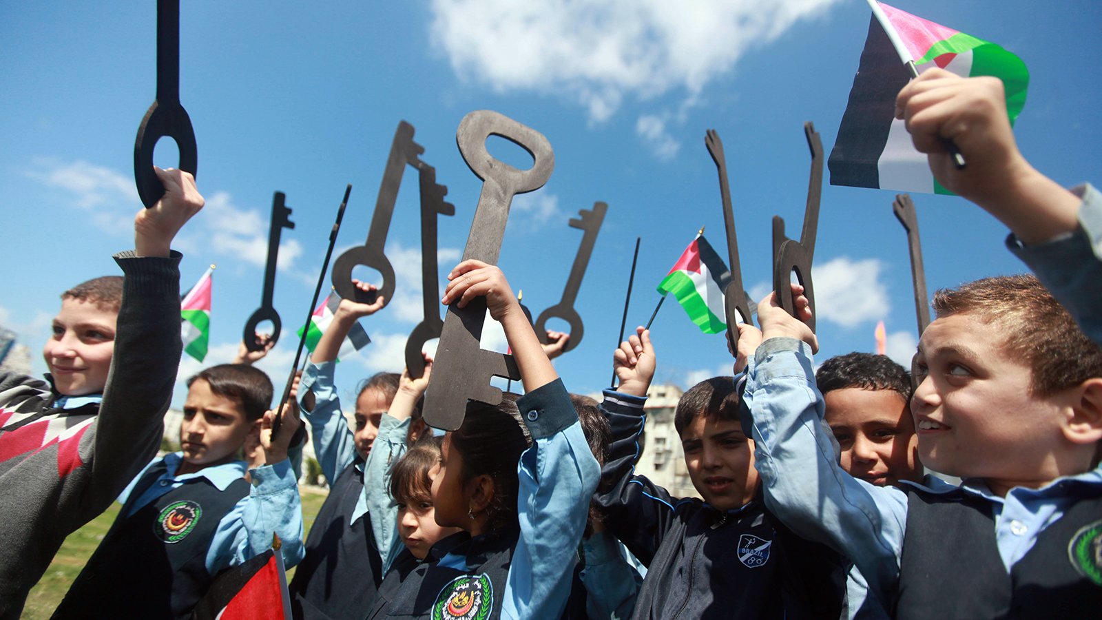 الصورة تظهر مجموعة من الأطفال يقفون في الهواء الطلق، يحملون مفاتيح كبيرة وأعلام فلسطينية. يبدو أنهم يعبرون عن تأييد لقضية معينة أو احتفال بذكرى تاريخية. الأطفال مبتسمون ومتحمسون، مما يعكس روح الفرح والاحتفاء والوعي بالقضايا المتعلقة بفلسطين. السماء صافية وملونة بالغيوم، مما يعطي شعوراً بالحيوية والنشاط.