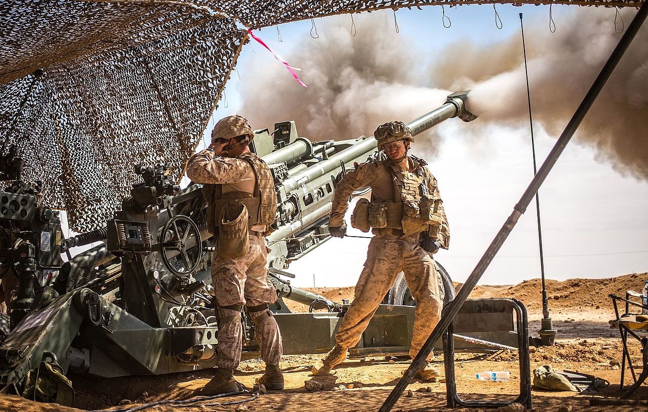 در این تصویر، دو سرباز در یک محیط نظامی دیده می‌شوند که به یک توپخانه بزرگ نزدیک هستند. یکی از سربازان در حال کار با ابزارهای مربوط به توپخانه است، در حالی که دیگری در حال نظارت بر عملیات به نظر می‌رسد. دود ناشی از شلیک گلوله به سمت آسمان می‌رود و پرده‌ای از توری برای حفاظت سربازان قرار داده شده است. فضای اطراف خشک و بیابانی به نظر می‌رسد، که نشان‌دهنده یک عملیات نظامی در یک منطقه جنگی است.