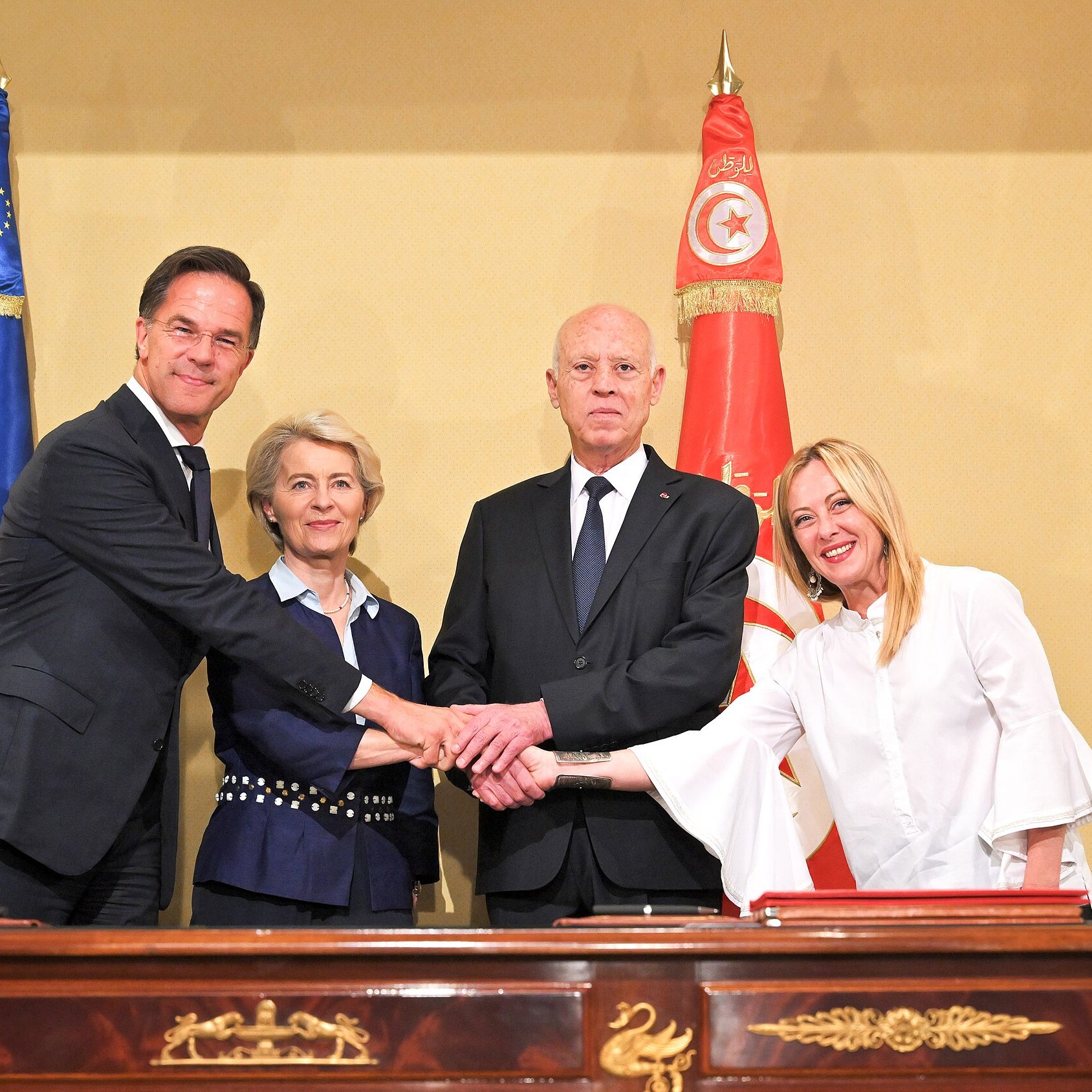 L'image montre quatre personnes se serrant la main autour d'une table. À gauche, un homme en costume sombre, probablement un représentant d'un pays européen. À sa droite, une femme avec des cheveux blonds peignés, semblant être une dirigeante politique. Au centre, un homme en costume sombre, certainement le président d'un pays, et à sa droite, une femme vêtue de blanc, probablement une autre dirigeante. En arrière-plan, on peut voir des drapeaux de l'Union européenne et de la Tunisie, ce qui suggère une rencontre diplomatique.