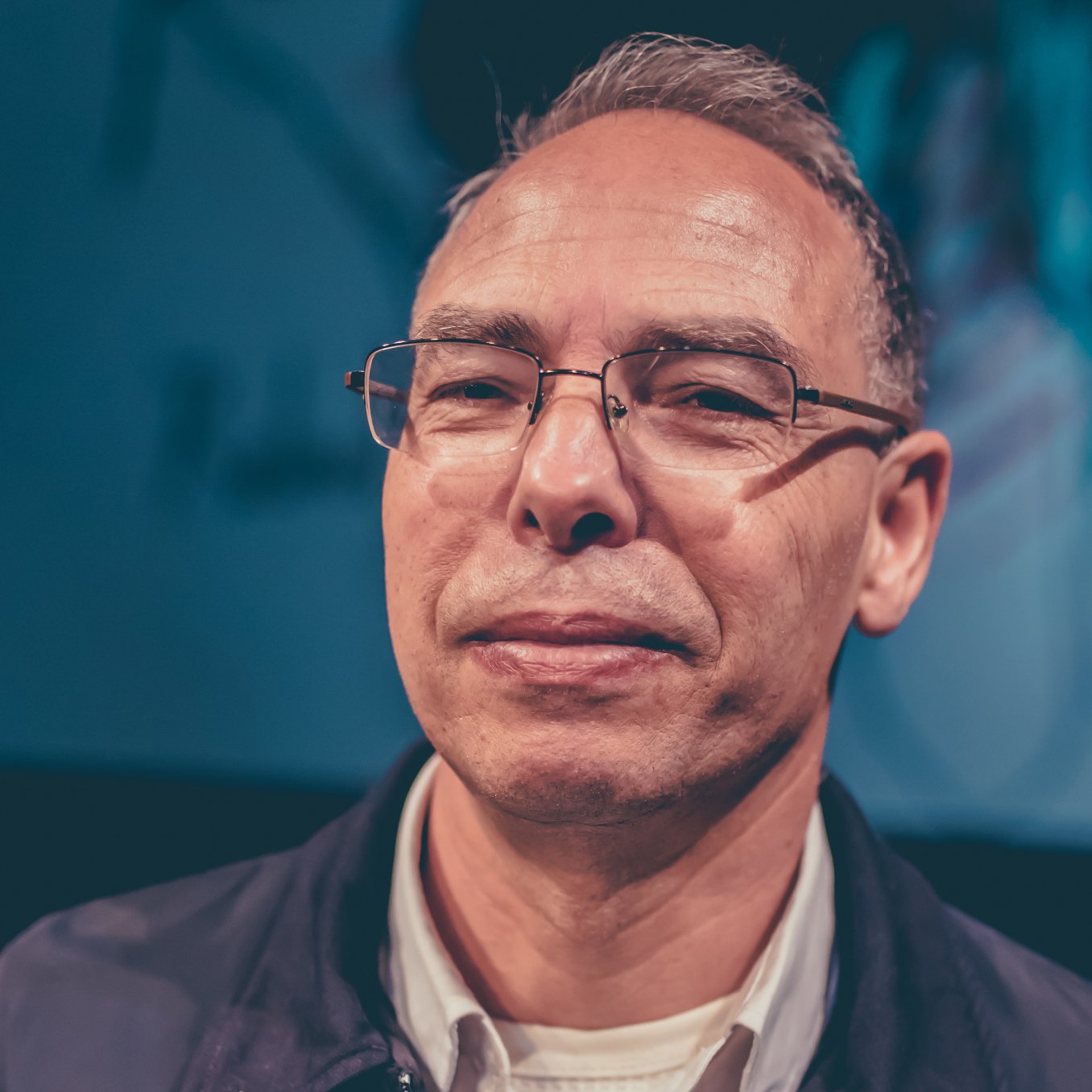 A middle-aged man with glasses and a slight smile, standing against a blurred background.