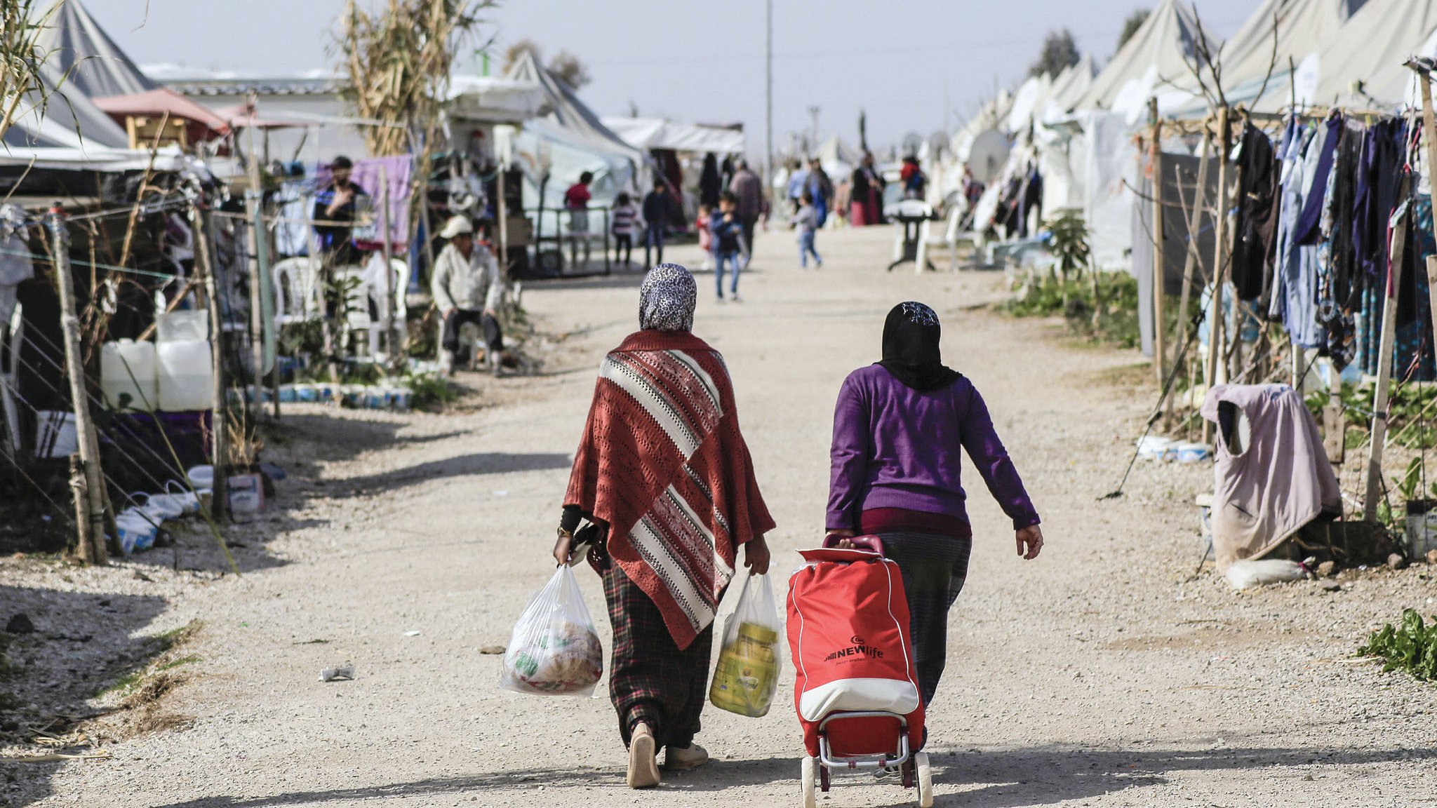 L'image montre une scène d'un camp, probablement un camp de réfugiés ou un site temporaire. Deux femmes marchent sur un chemin de terre, portant des sacs. L'une d'elles pousse une poussette. En arrière-plan, on aperçoit des tentes et des structures improvisées, avec d'autres personnes visibles dans l'environnement. L'atmosphère semble être celle d'une communauté, avec des éléments de vie quotidienne.