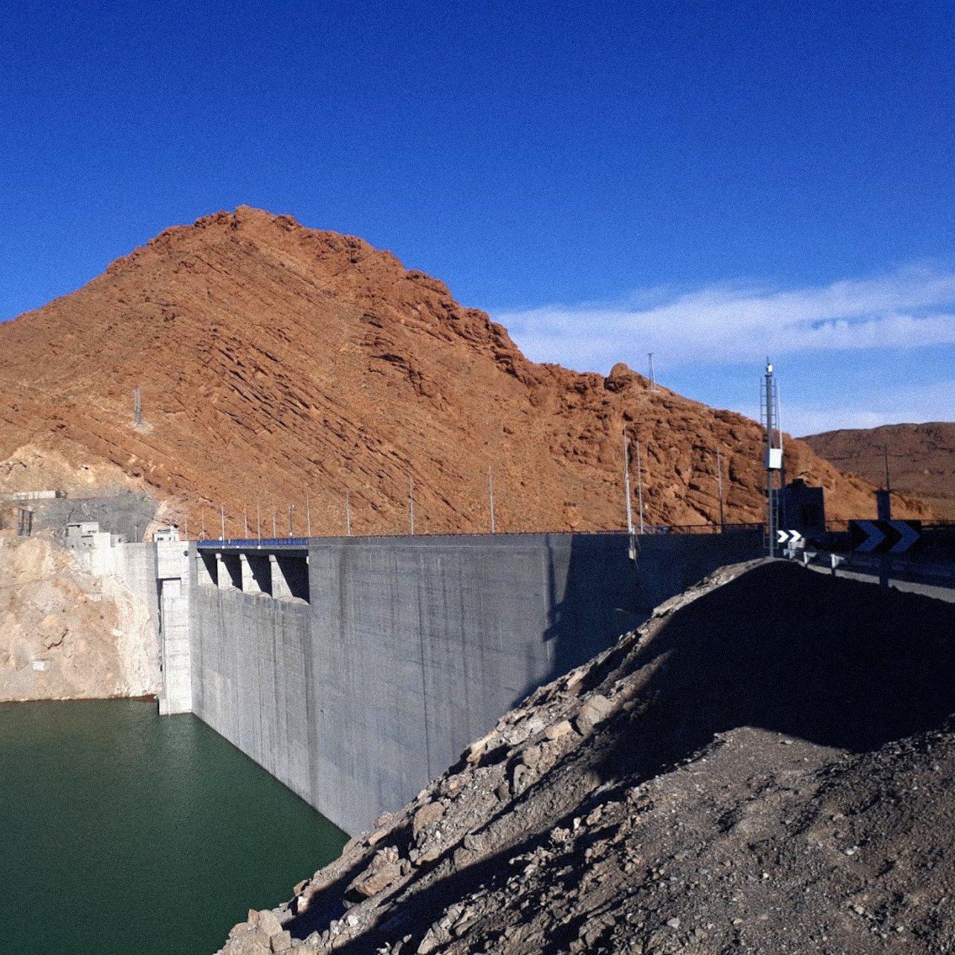 La imagen muestra un gran embalse o represa construida de concreto, situada a la orilla de un cuerpo de agua de color verde. Detrás de la represa se puede ver una montaña de tonalidades marrones y naranjas, con un cielo despejado de un azul brillante en el fondo. La estructura de la represa es sólida y parece estar bien mantenida, mientras que el terreno alrededor tiene un aspecto árido y rocoso.