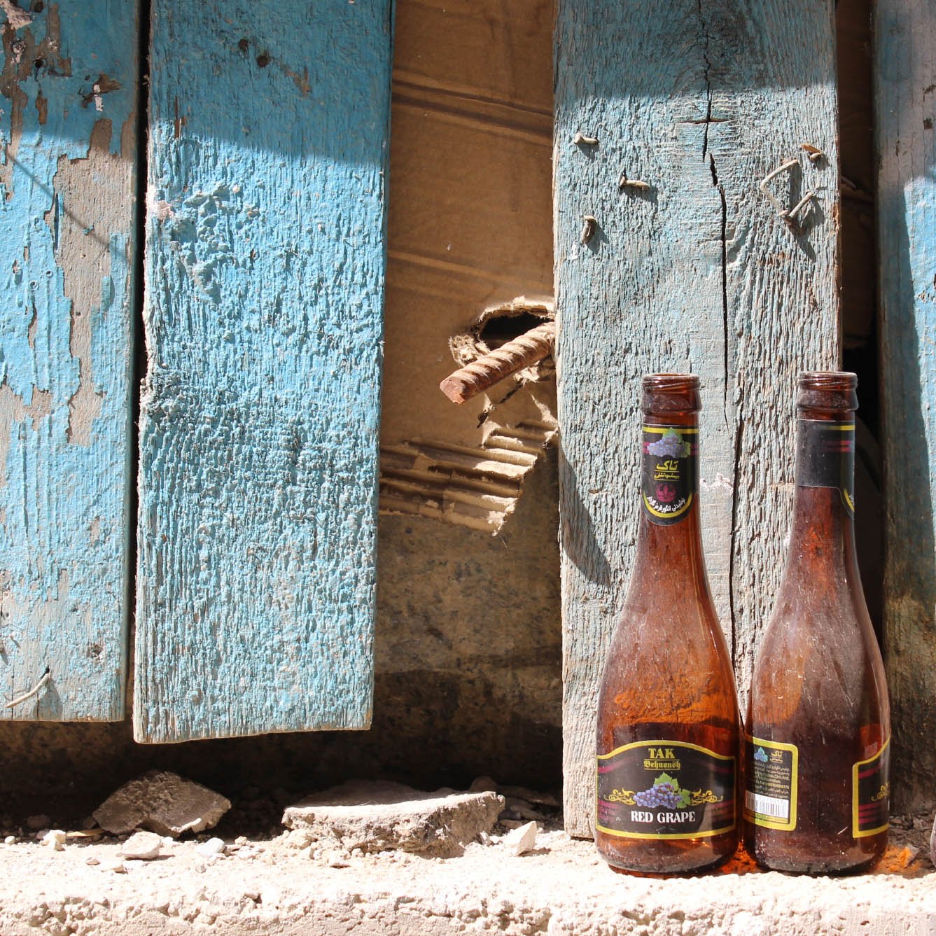 در این تصویر، سه بطری خالی در کنار دیواری که رنگ آبی دارد و کمی فرسوده به نظر می‌رسد، قرار گرفته‌اند. دیواره دارای نواحی ترک‌خورده و برخی زواید و نقص‌هاست. بطری‌ها به صورت نامرتب در کنار هم قرار دارند و بافتی کهنه و رنگ‌هایی مات دارند، که به تصویر حس قدیمی و رها شده‌ای می‌بخشد. در پس‌زمینه، تکه‌هایی از دیوار نیز قابل مشاهده است.