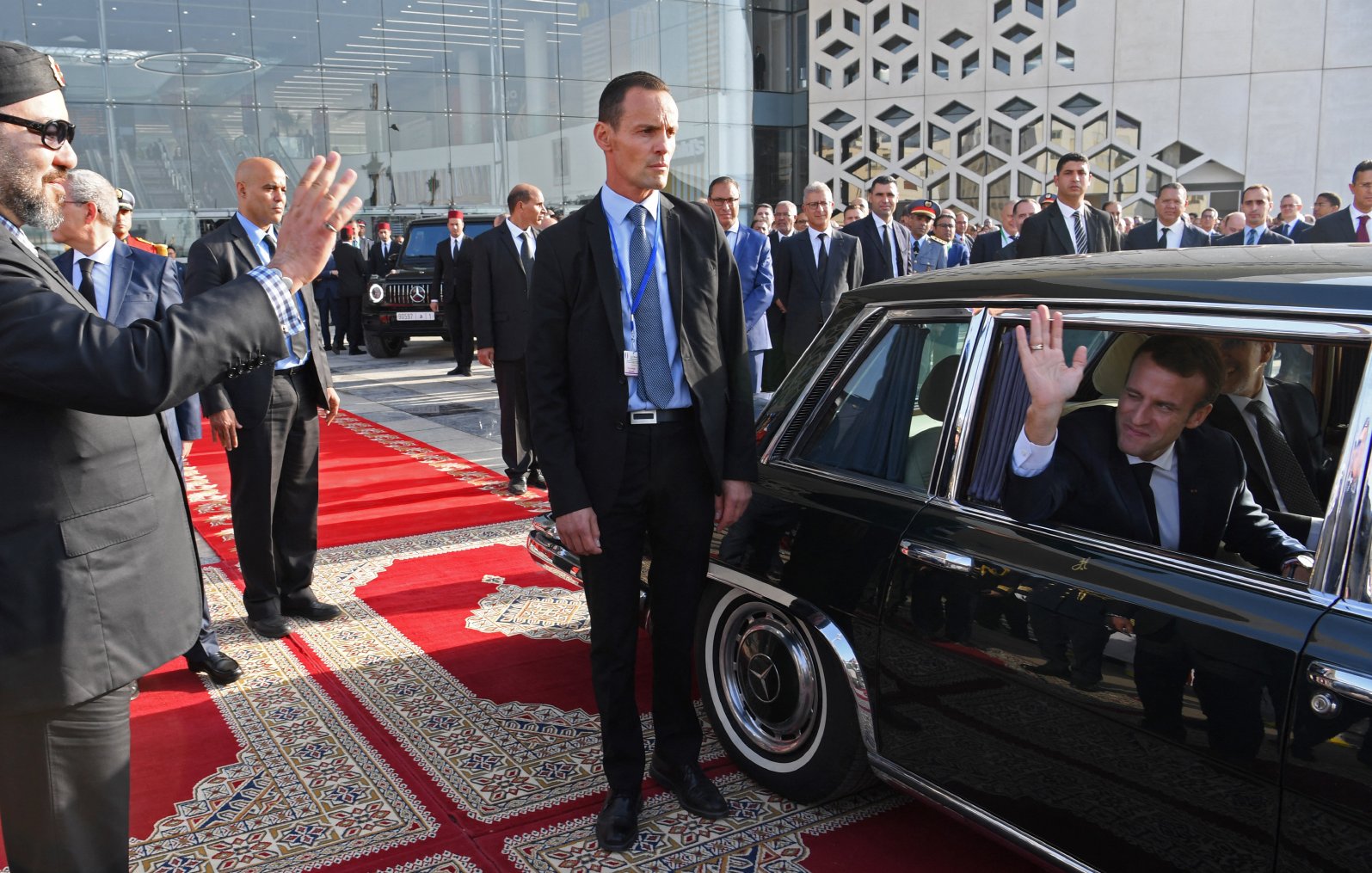 L'image montre une scène d'accueil officielle. On y voit un homme portant des lunettes de soleil et un chapeau, qui salue avec la main. À sa droite, un homme en costume, apparemment en position de sécurité, observe la situation. De l'autre côté, un homme assis dans une voiture noire, qui semble répondre au salut en levant la main. Le décor est élégant, avec un tapis rouge et des bâtiments modernes en arrière-plan.