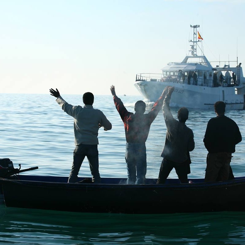 The image depicts four individuals in a small boat on calm waters, facing away from the camera. They are raising their arms, seemingly in a gesture of celebration or greeting. In the background, a larger boat is approaching, with several people visible on it. The scene is set against a clear sky, suggesting a peaceful day on the water.