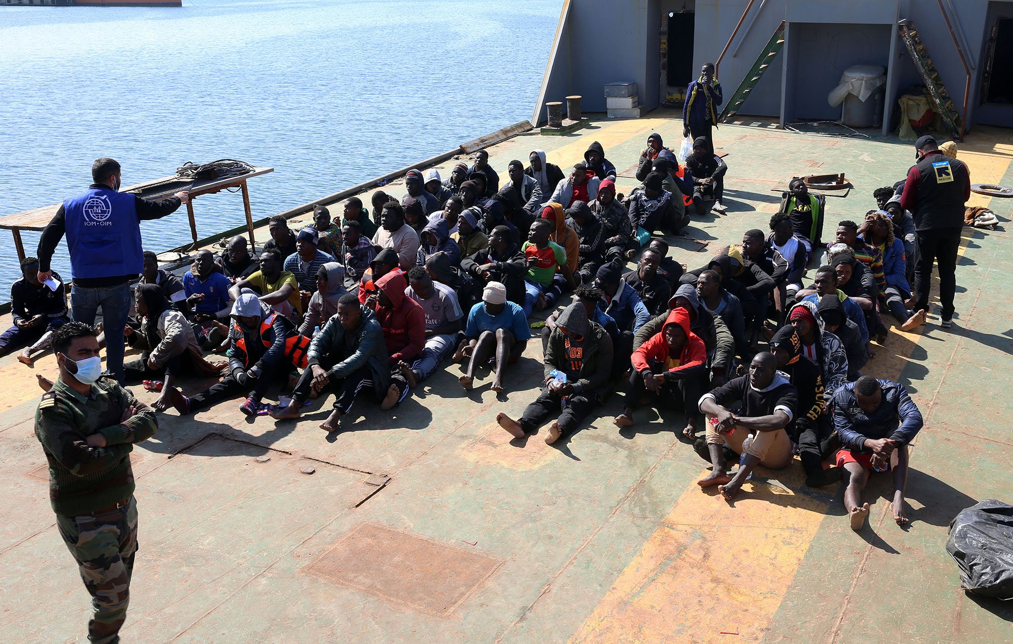 در این تصویر، گروهی از افراد در یک کشتی نشسته‌اند. آنها در حالتی متراکم و منظم روی عرشه نشسته‌اند و بیشتر آنها لباس‌های گرم به تن دارند. برخی از آنها هم کلاه و ماسک به صورت دارند. در سمت چپ تصویر، فردی با یونیفورم نظامی و شخصی دیگر با جلیقه آبی در حال نظارت بر این گروه هستند. در پس‌زمینه، آب دریا و بخشی از ساحل قابل مشاهده است. جو کلی تصویر به نظر می‌رسد که مربوط به یک عملیات نجات یا مهاجرت باشد.