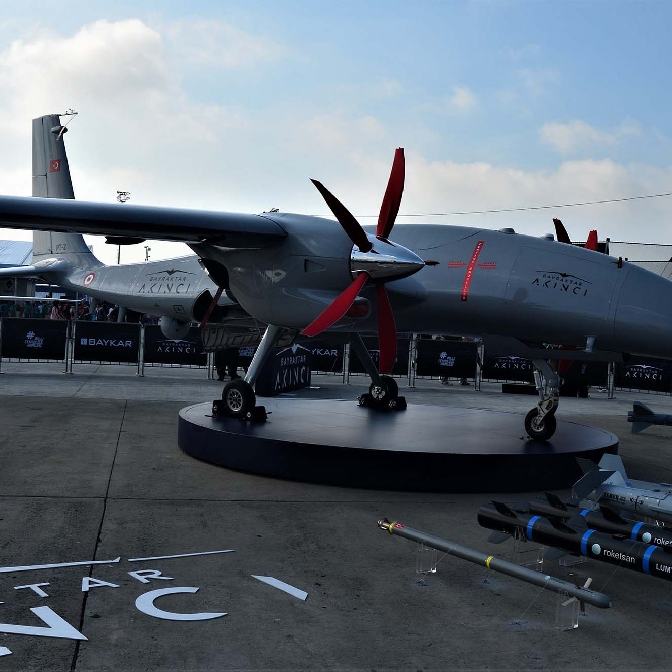 La imagen muestra un dron militar de la marca Baykar. Está expuesto en un evento, con un diseño aerodinámico y detalles en rojo. El dron tiene dos hélices rojas en el frente y un fuselaje gris, apoyado sobre una plataforma giratoria. Al fondo, se pueden observar otros drones y un ambiente que sugiere un evento de exhibición o industria militar. También hay misiles en primer plano, lo que indica su capacidad bélica.