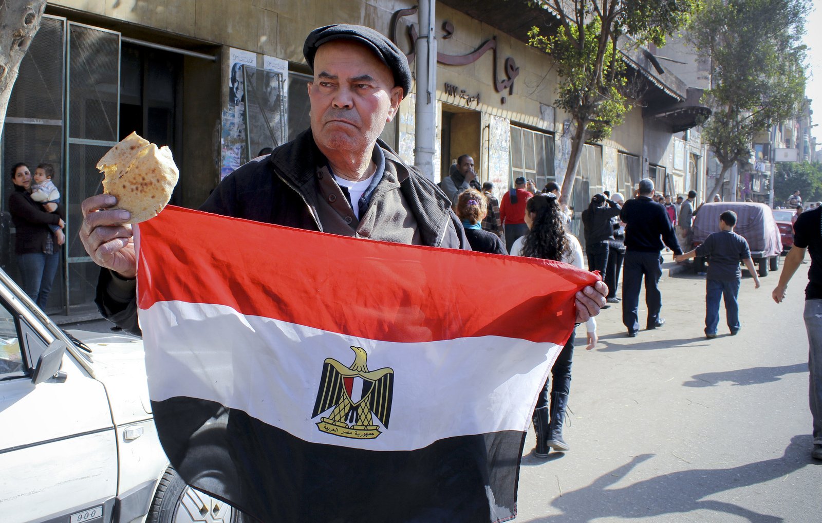 L'image montre un homme tenant un drapeau égyptien, avec le symbole de l'Égypte au centre. Il tient également un morceau de pain dans l'autre main. Demain la ville est animée, avec plusieurs personnes en arrière-plan, ce qui suggère une atmosphère de rassemblement ou de manifestation. L'homme semble sérieux et déterminé, exprimant un fort sentiment national. L'environnement urbain, avec des bâtiments et des passants, reflète la vie quotidienne en Égypte.