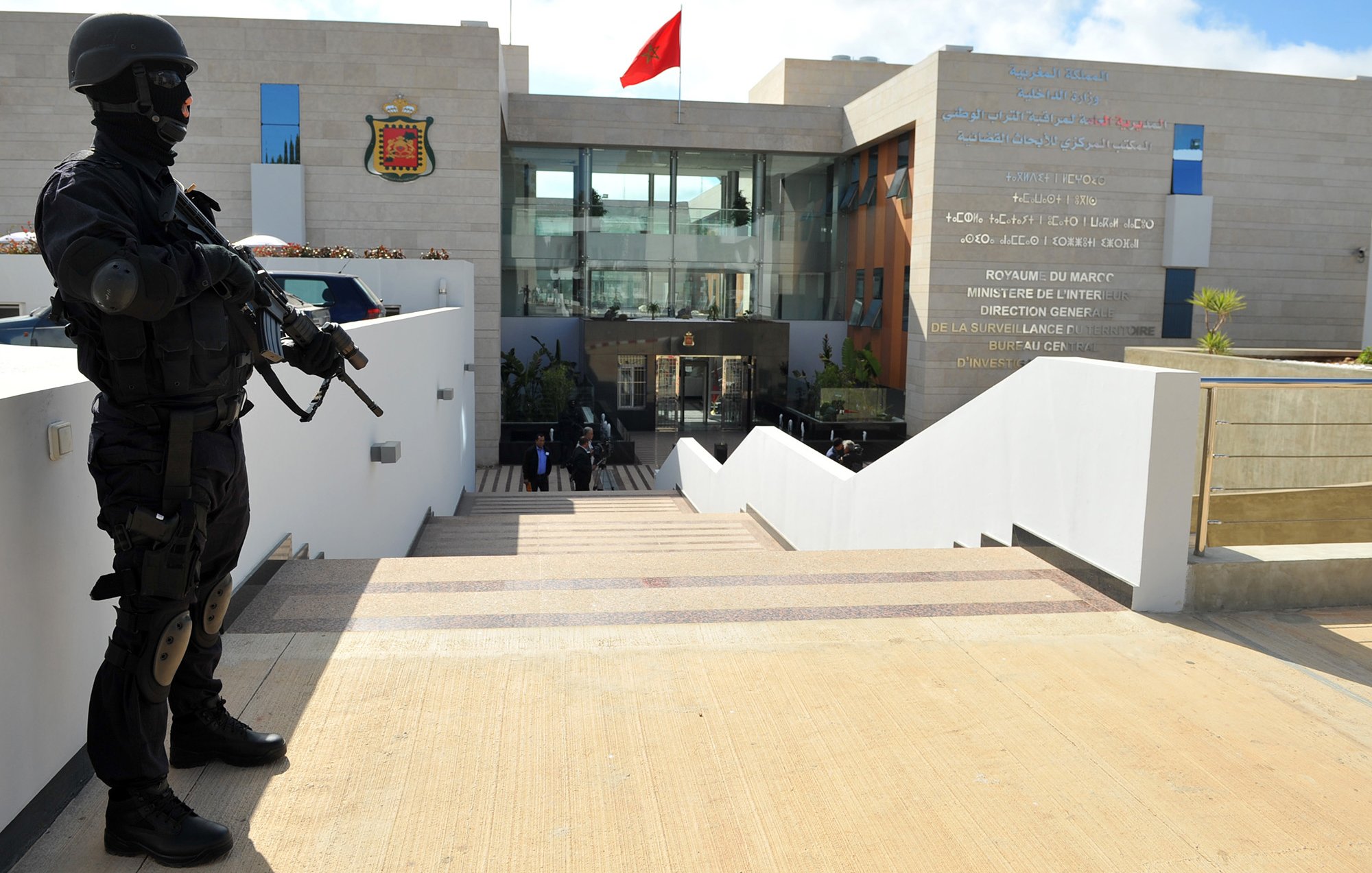 The image depicts a heavily armed police officer or security personnel standing at attention outside a building. The officer is dressed in black tactical gear and is holding a firearm. In the background, there is a modern structure with a large entrance, and the Moroccan flag is prominently displayed on top. The building appears to be a government facility, likely related to security or law enforcement, as indicated by the presence of the armed guard. The setting features stairs leading up to the entrance, with a clean, contemporary design.