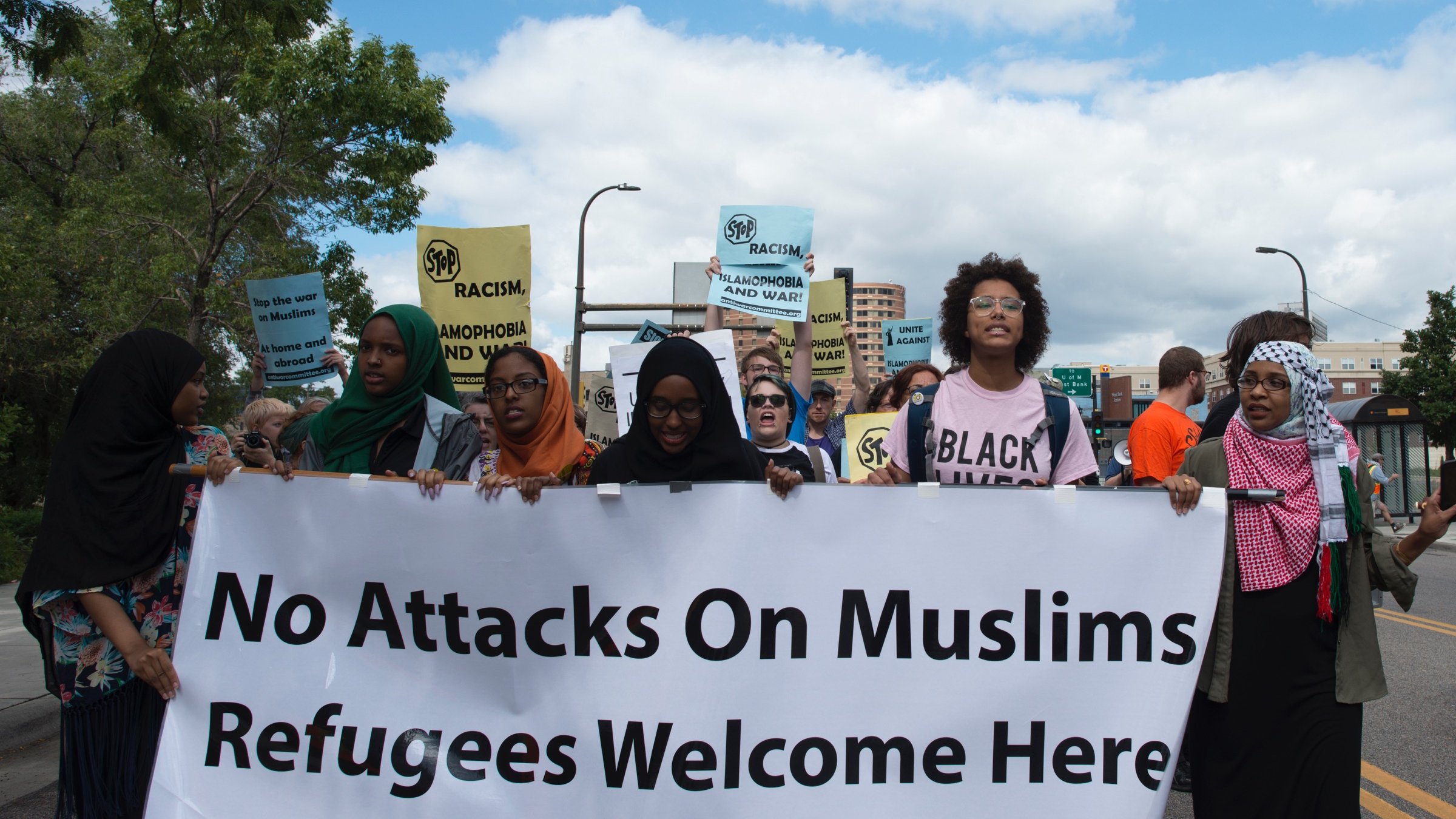 L'image montre un groupe de manifestants en train de marcher dans une rue. Ils portent des pancartes avec des messages tels que "No Attacks On Muslims" et "Refugees Welcome Here". Les participants, majoritairement de culture musulmane, affichent des expressions déterminées. Le ciel est partiellement nuageux, et on peut voir des bâtiments en arrière-plan. L'atmosphère semble engager et militante, soulignant des thèmes de solidarité et de droits humains.
